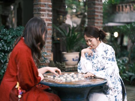 Two women in traditional attire focus on a board game outdoors, surrounded by greenery.