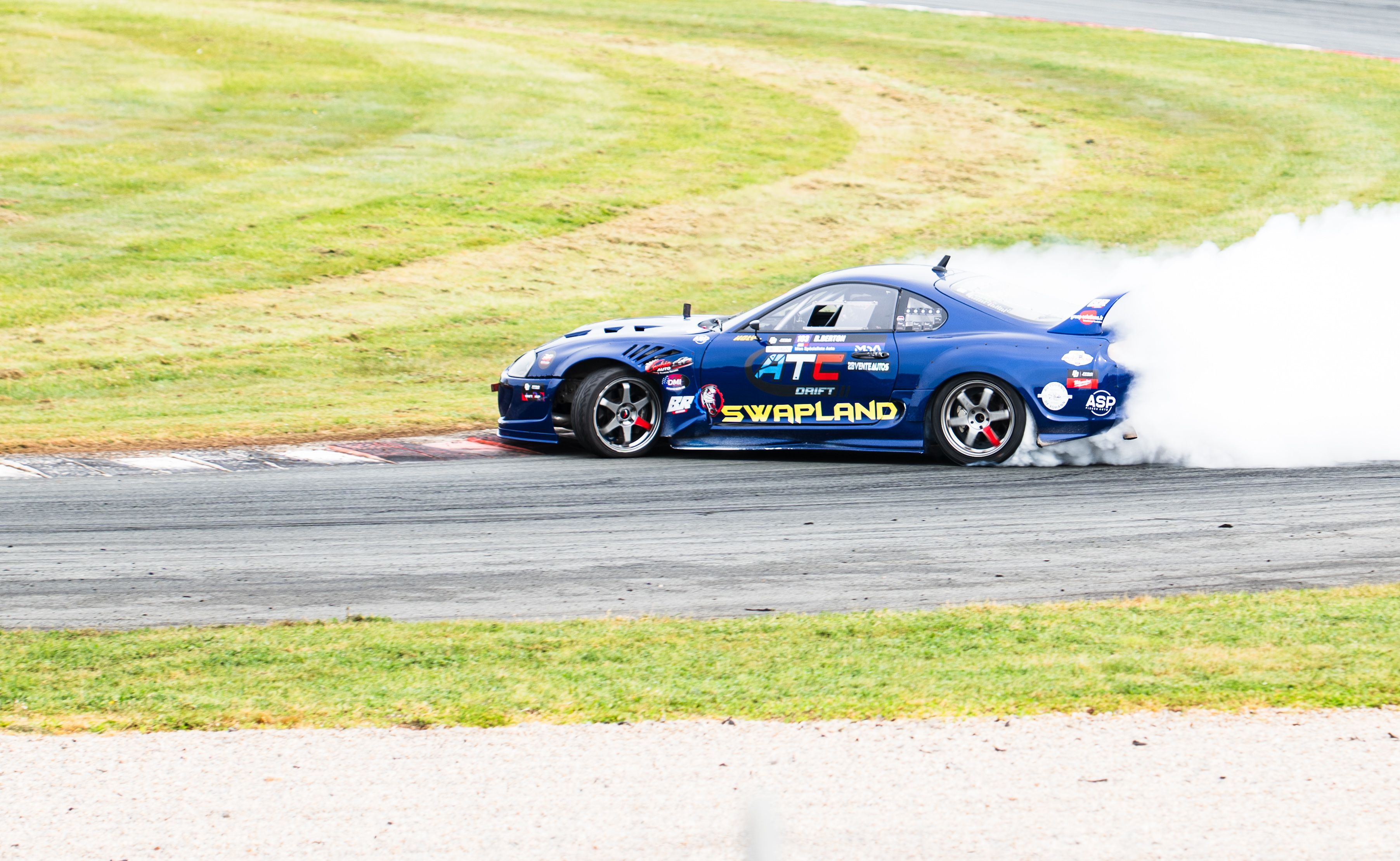 A blue racing car drifting on a track, creating a smoke trail under sunny conditions.