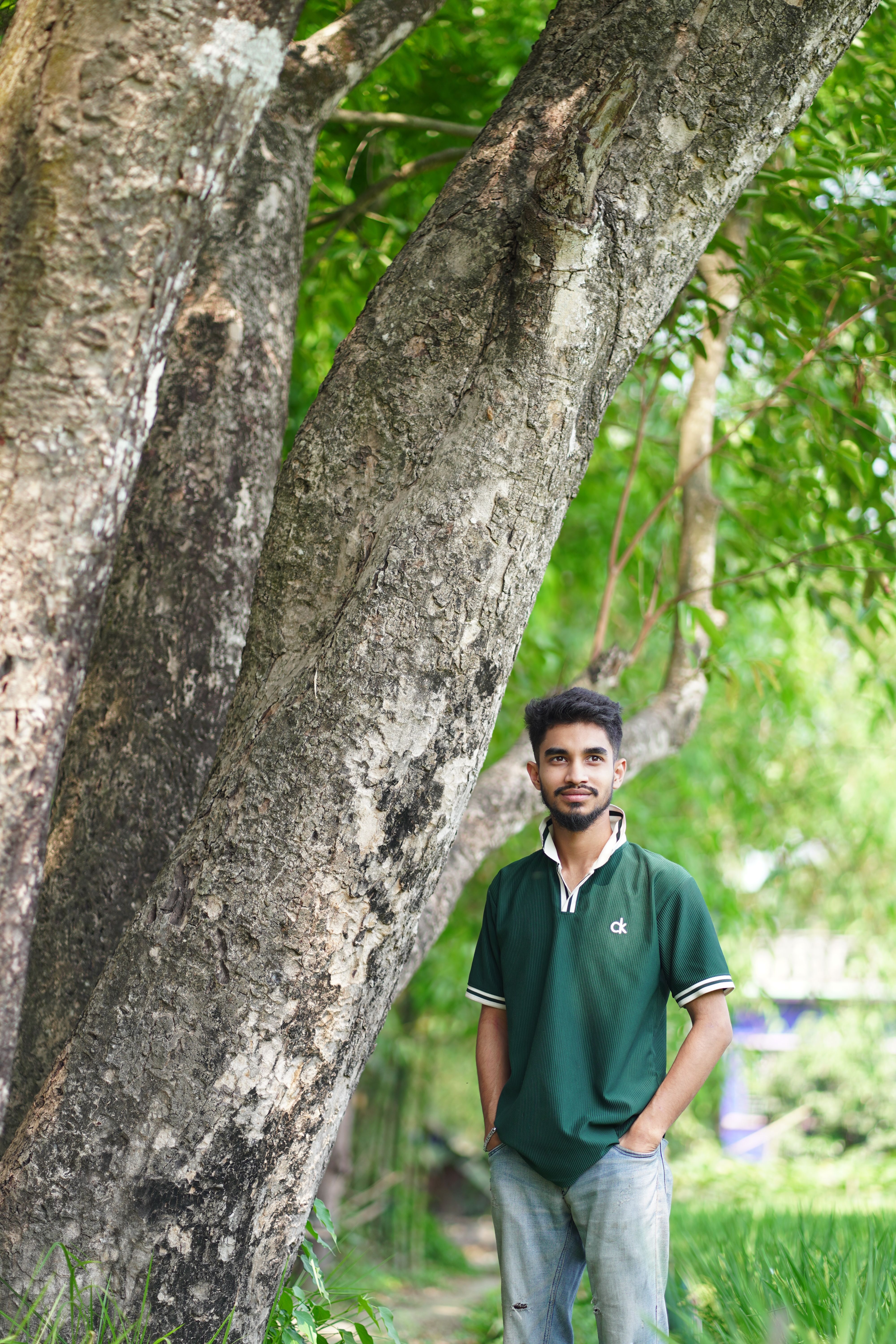 Portrait of a young man standing beside a large tree in a green outdoor setting.