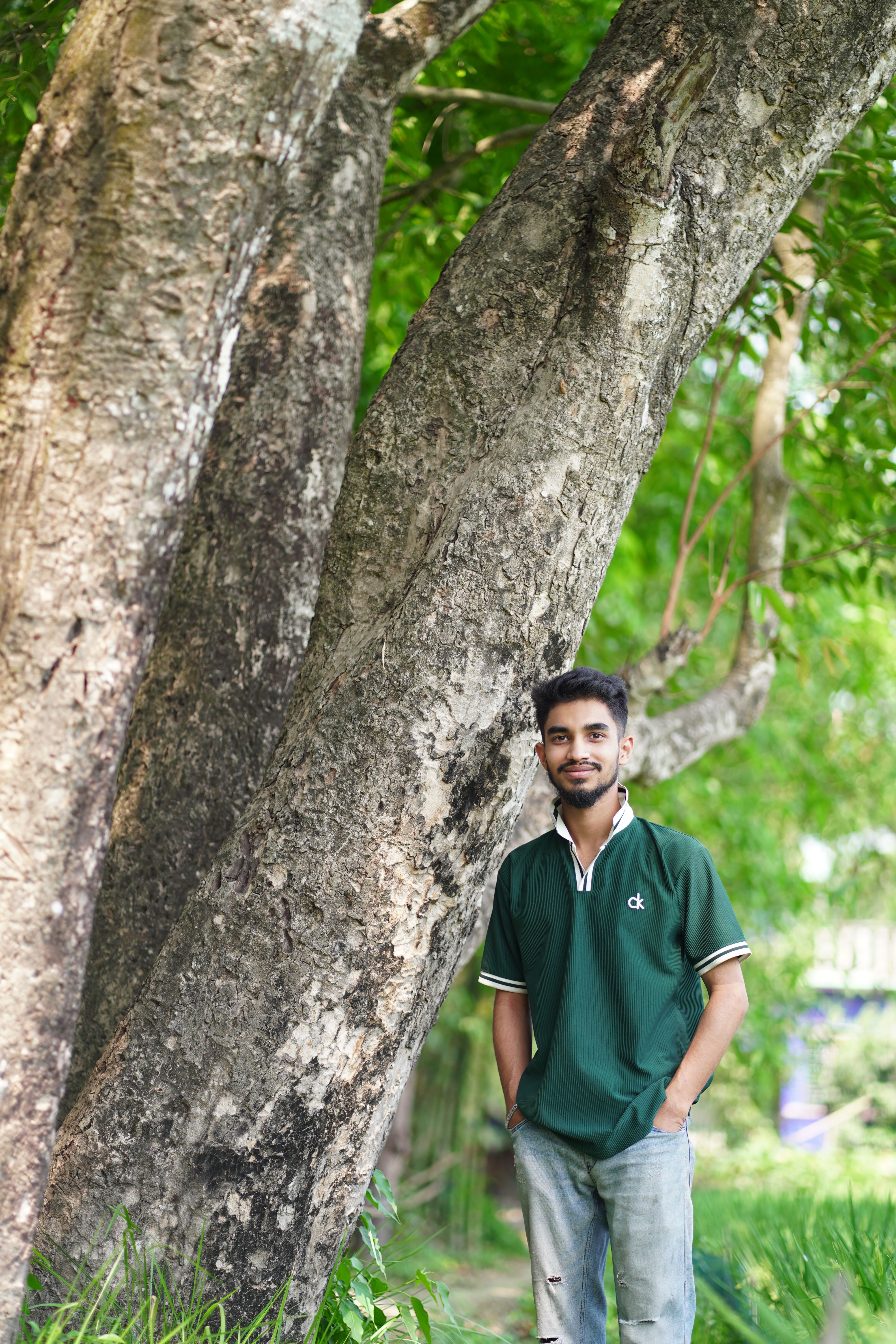 A young man stands by a large tree in a tranquil outdoor setting. Greenery surrounds.