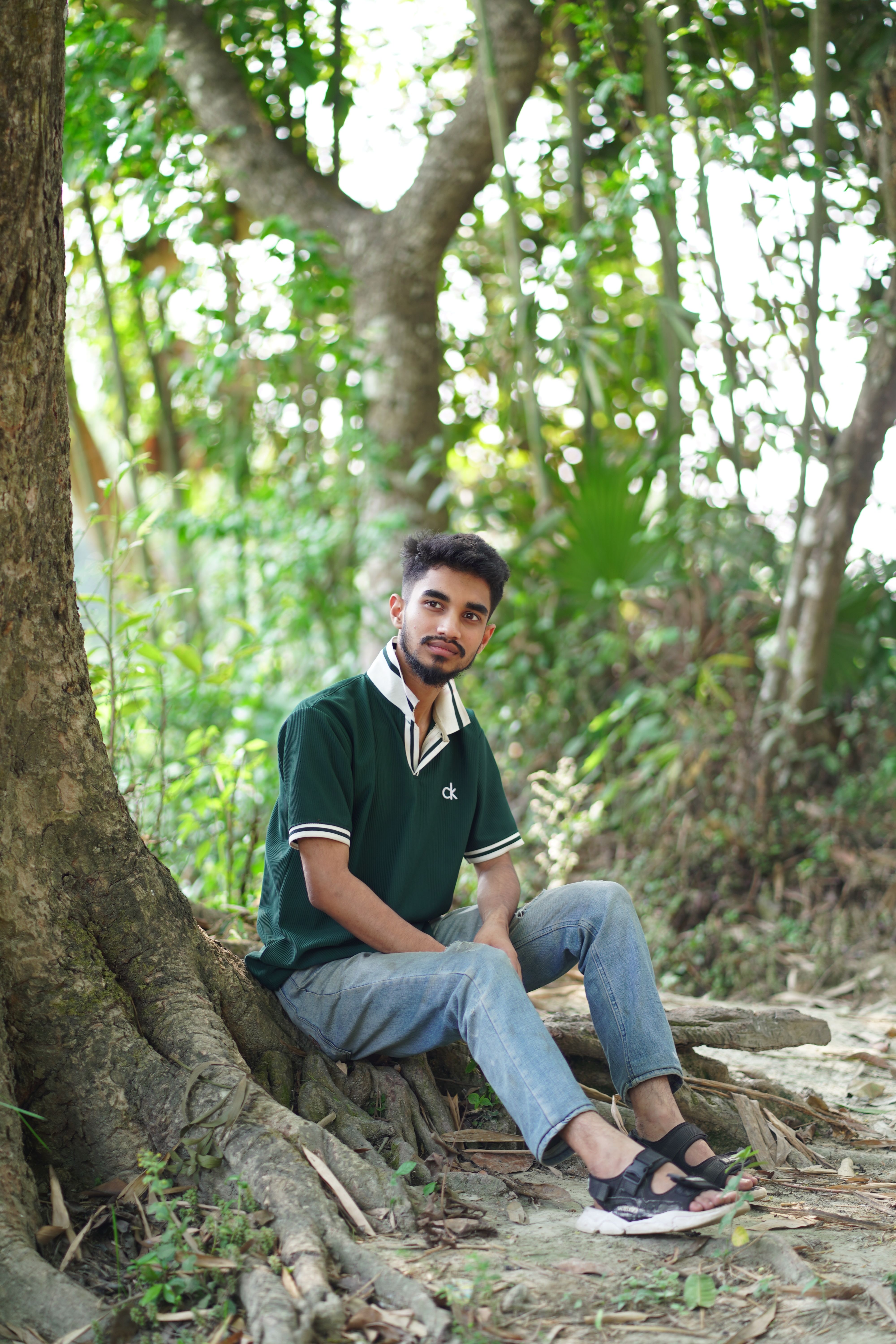 Casual portrait of a young man seated by a tree in a serene forest environment.