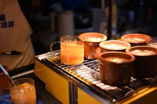 A vibrant street food stall showcasing steaming hot beverages in mugs at night, with warm lighting and an apron-clad vendor.