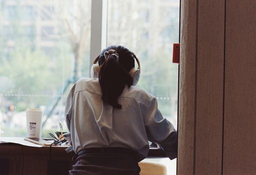 A woman wearing headphones studies by a window in a Beijing cafe, capturing a tranquil and focused moment.