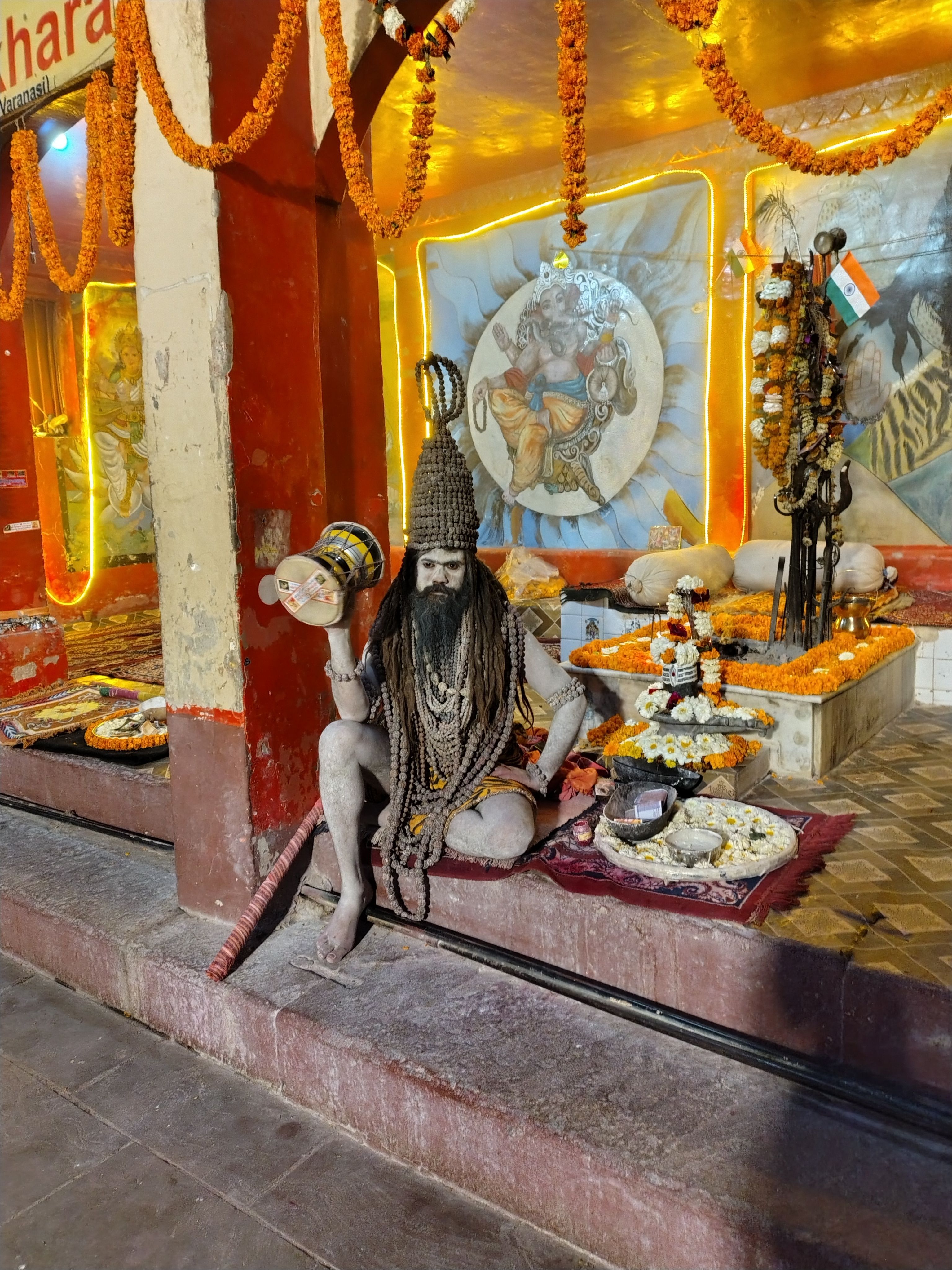 A holy man with intricate decorations sits at an Indian shrine, surrounded by offerings and bright colors.