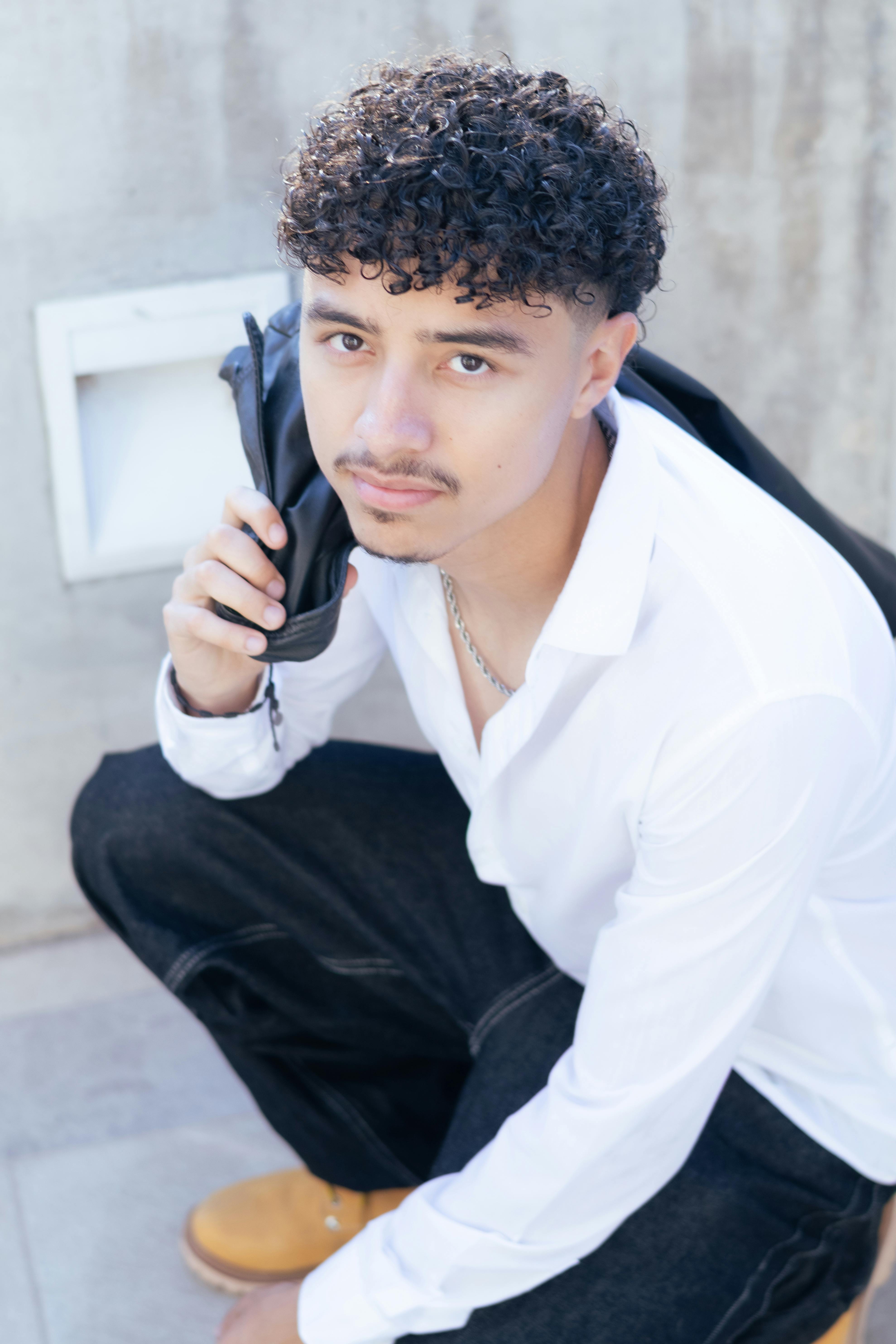 Stylish young man in urban attire seated outdoors in Aguascalientes, displaying modern fashion.