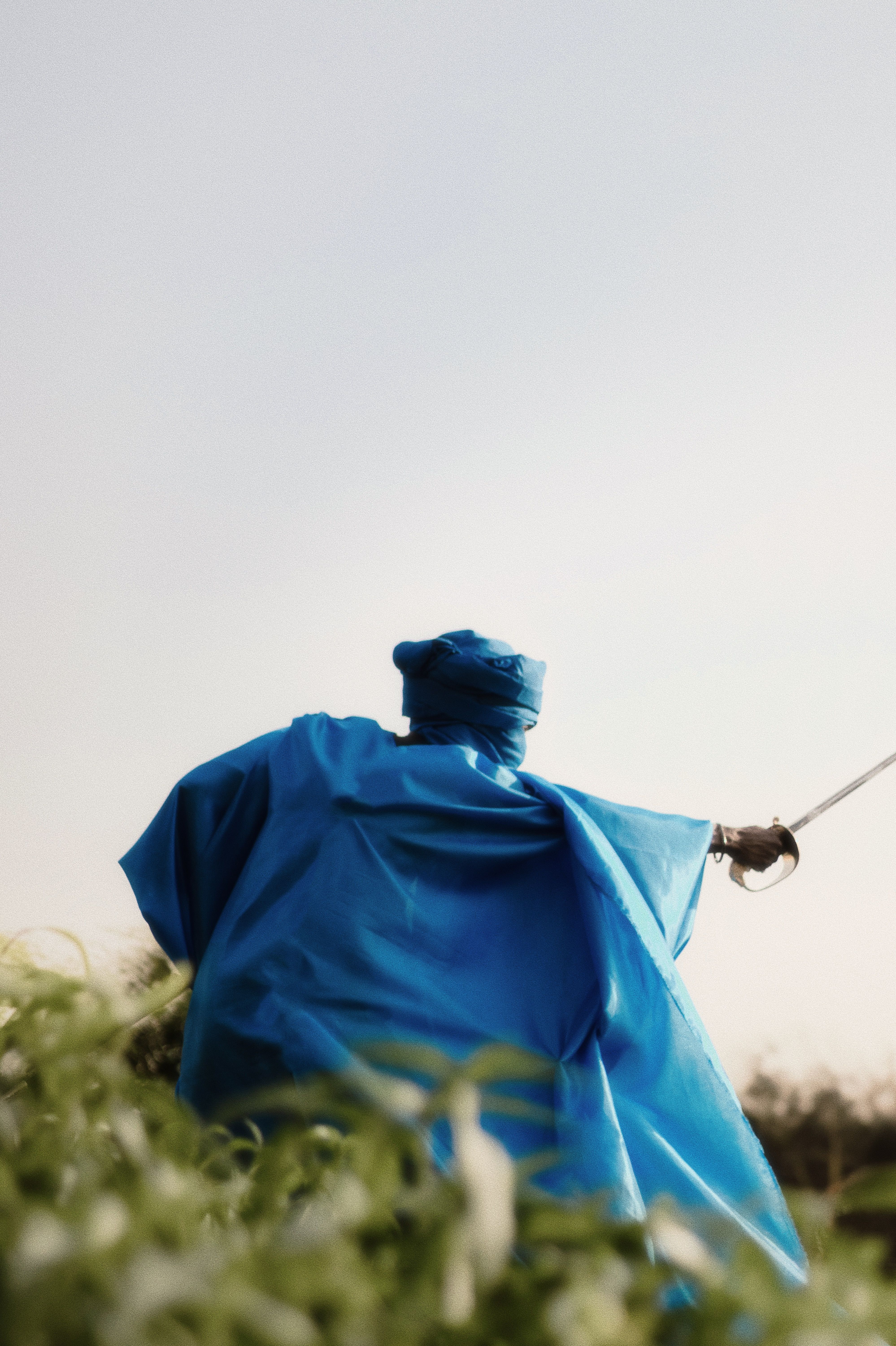 A man in a vibrant blue traditional robe holds a sword outdoors, exuding strength and cultural heritage.