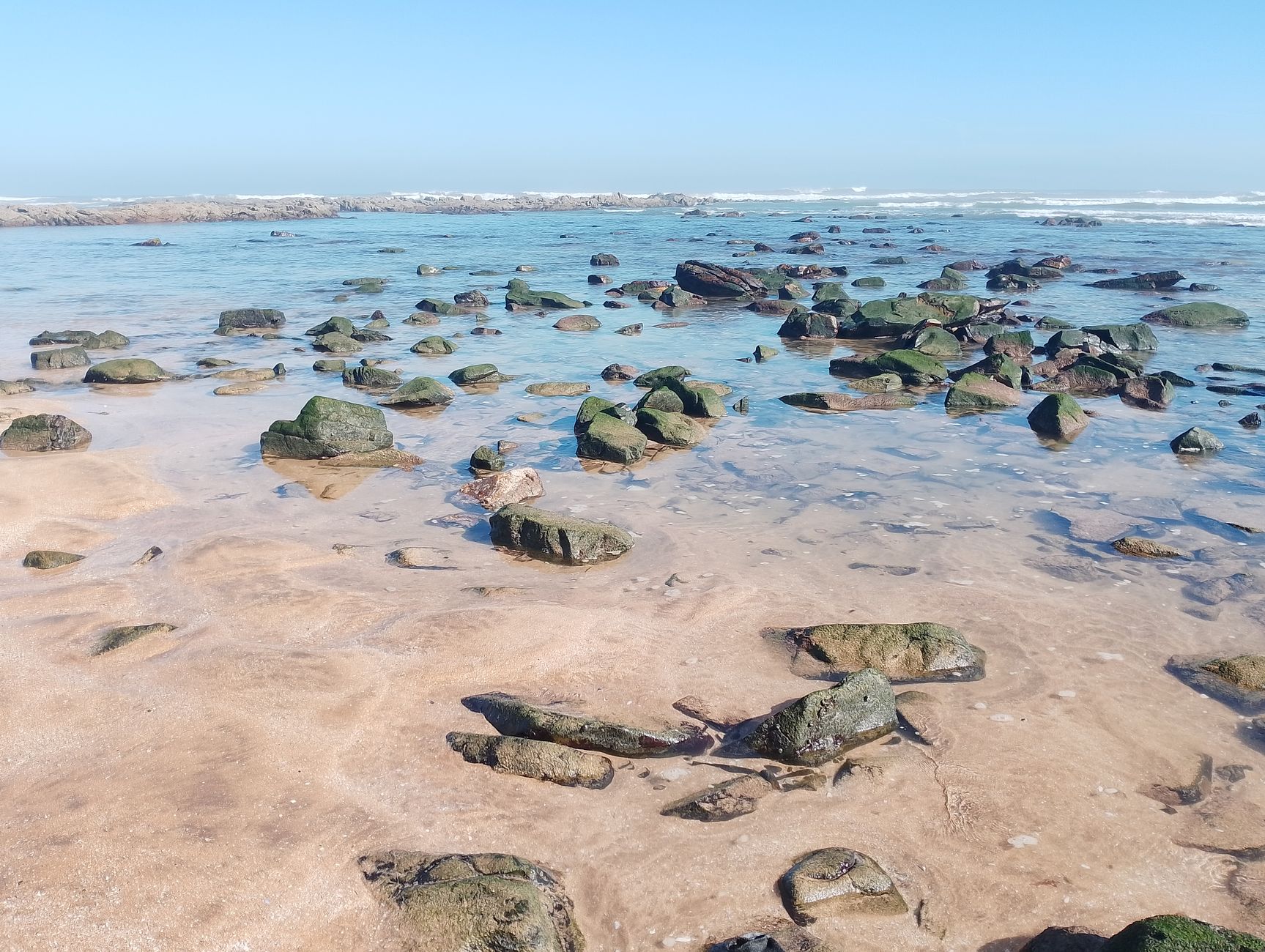 Explora una serena costa rocosa con marea baja, arena suave y olas suaves bajo un cielo azul claro.