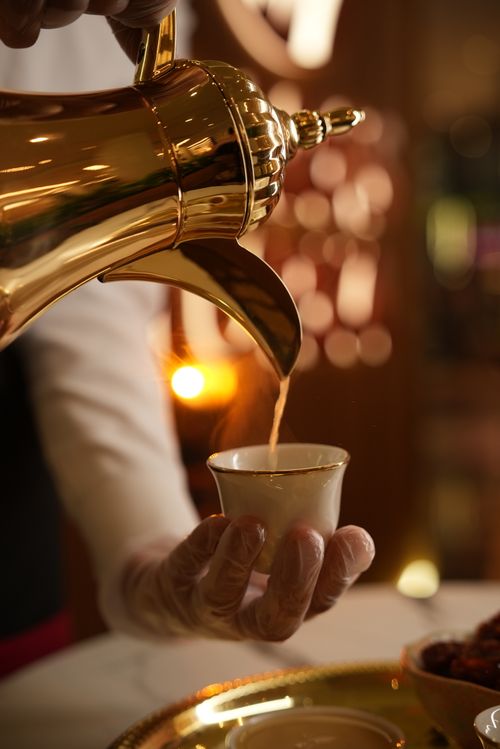 Free A gloved hand elegantly pours Arabic coffee from a golden pot into a traditional cup. Stock Photo