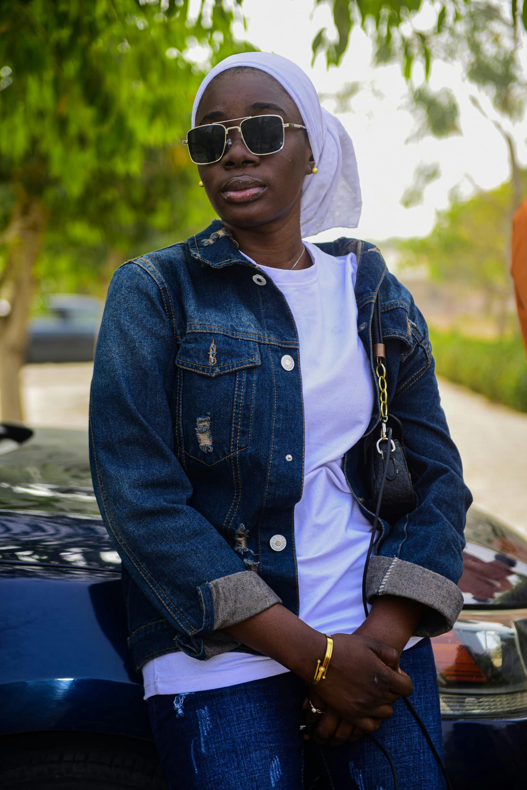 Fashionable woman in denim jacket and sunglasses leaning casually on a car outdoors.