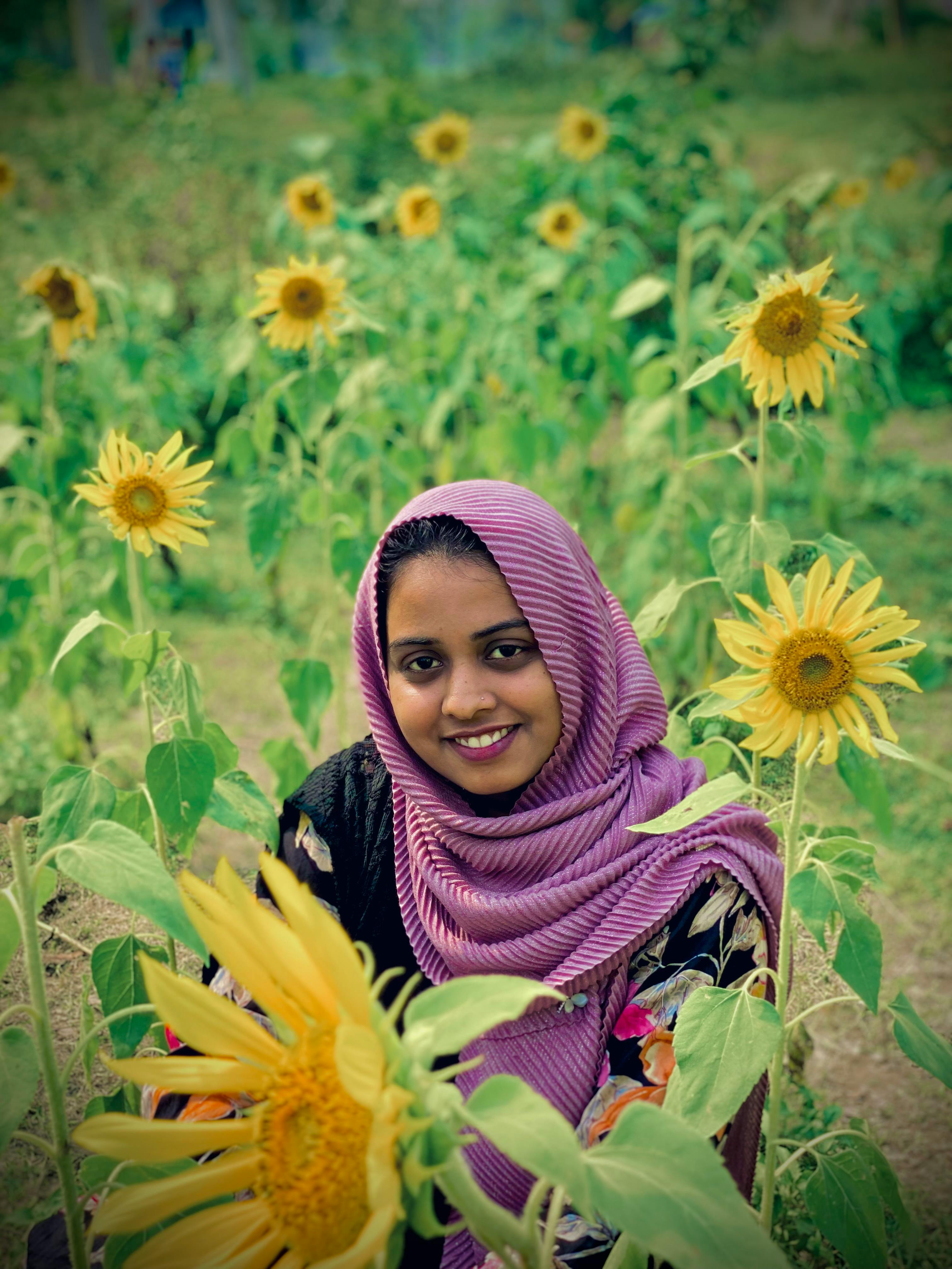 A young woman wearing a hijab smiles surrounded by blooming sunflowers in a vibrant garden.
