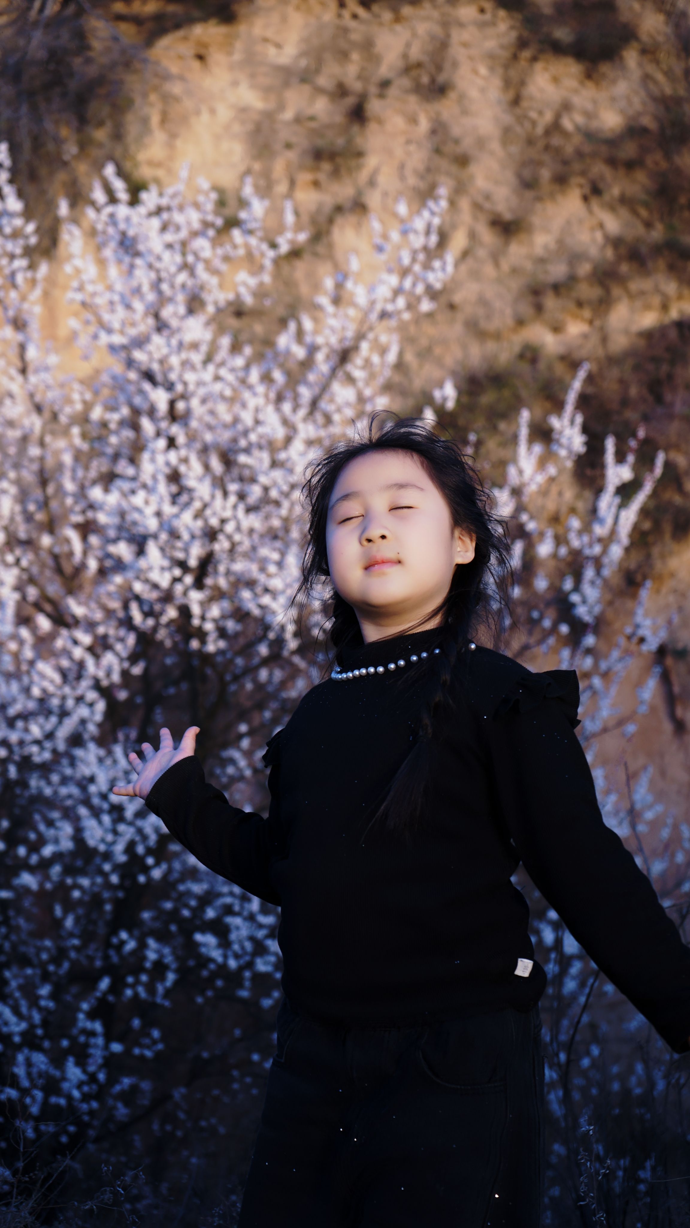 A young girl with closed eyes stands in front of blooming trees, embracing nature's serenity.