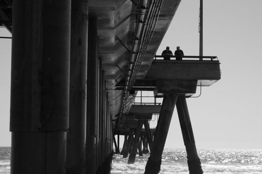 Artistic black and white image of a pier over the ocean with two people enjoying the view.