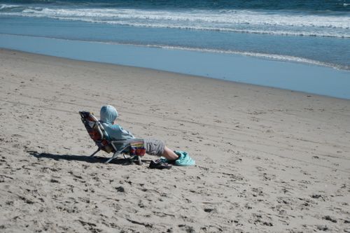 Free A person in a hoodie sits on a beach chair by the ocean, enjoying a sunny day. Stock Photo