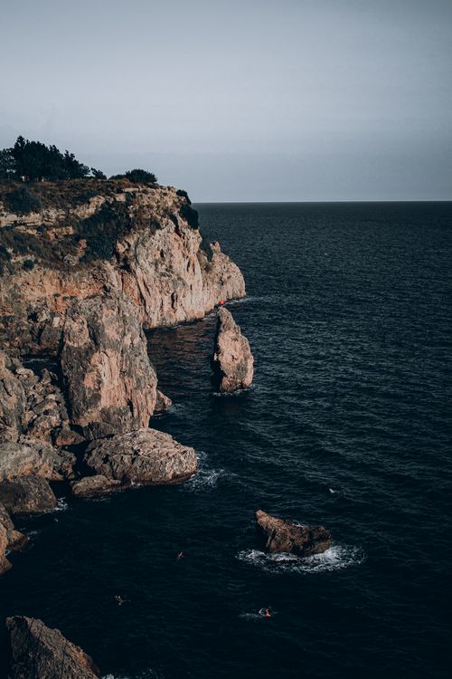 Free Rugged cliffside with deep blue ocean waves and distant swimmers at twilight. Stock Photo