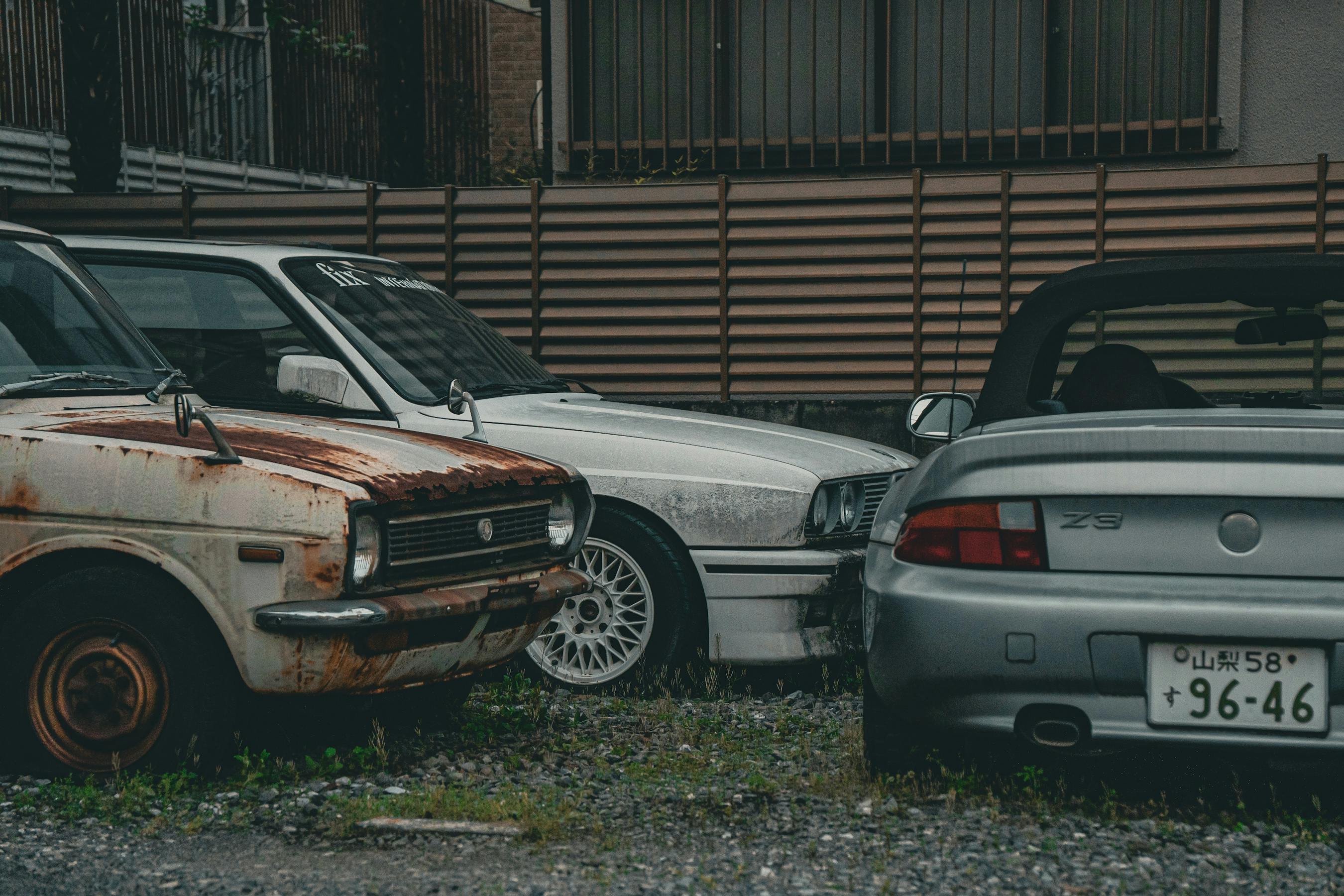 A rusted vintage car alongside modern vehicles in a gritty urban backdrop, highlighting contrast.