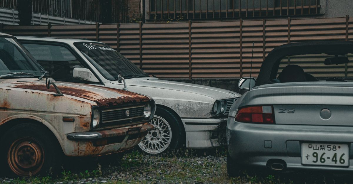 A rusted vintage car alongside modern vehicles in a gritty urban backdrop, highlighting contrast.