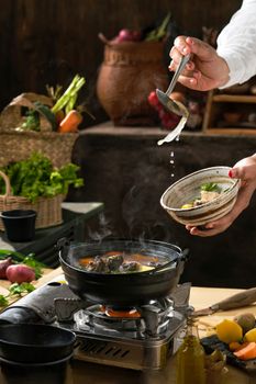 A chef's hand serving hot noodle soup from a pot, surrounded by fresh ingredients in a kitchen setting.