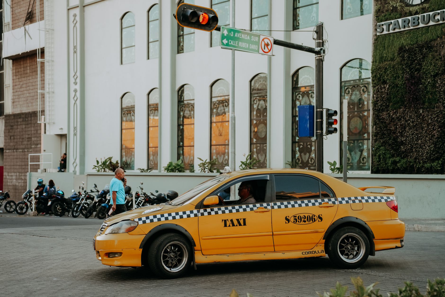 A bustling street scene in El Salvador featuring a yellow taxi and pedestrians on a busy intersection.