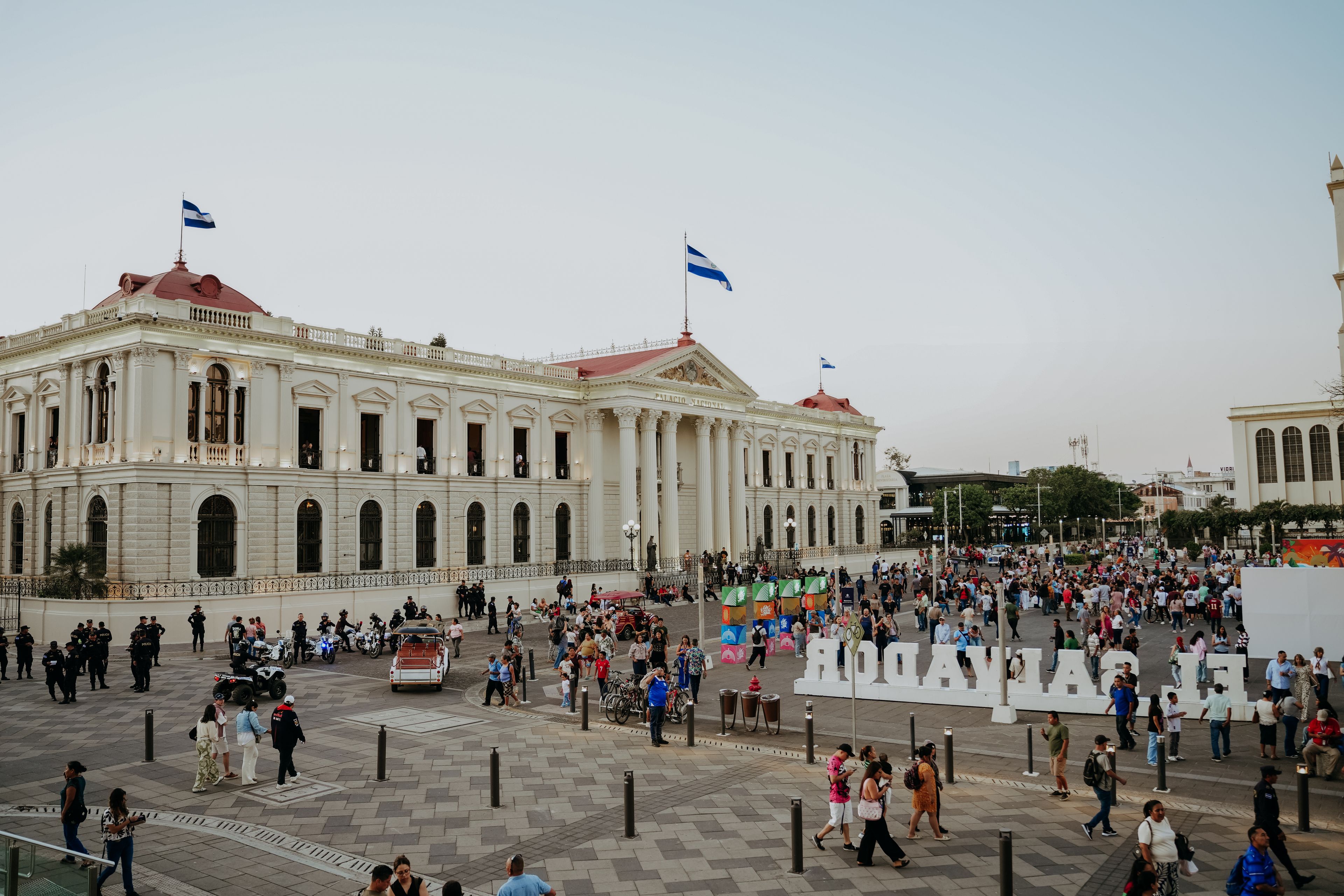 Crowded plaza scene in San Salvador with flags and historic buildings under a clear sky.