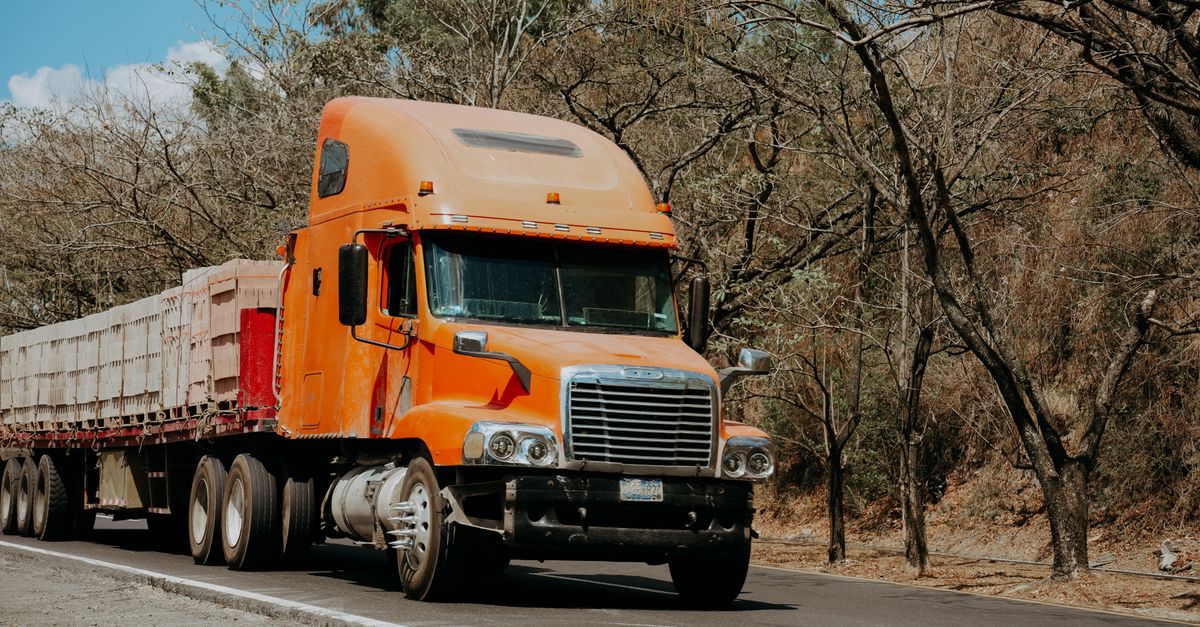 A vibrant orange Freightliner truck drives on a highway through a dry, scenic landscape in El Salvador.