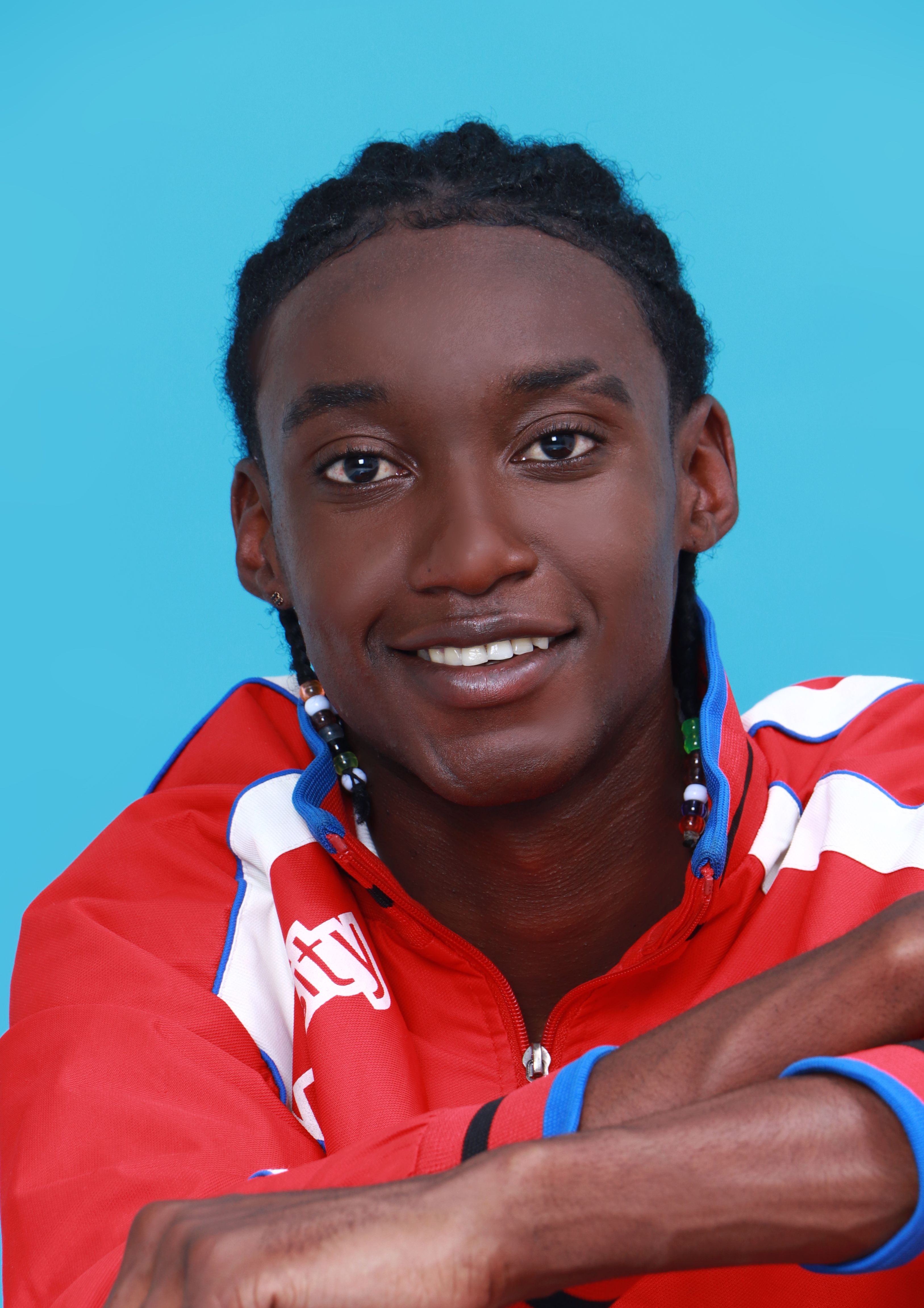 A cheerful young man with braided hair in a vibrant red jacket smiles confidently at the camera.