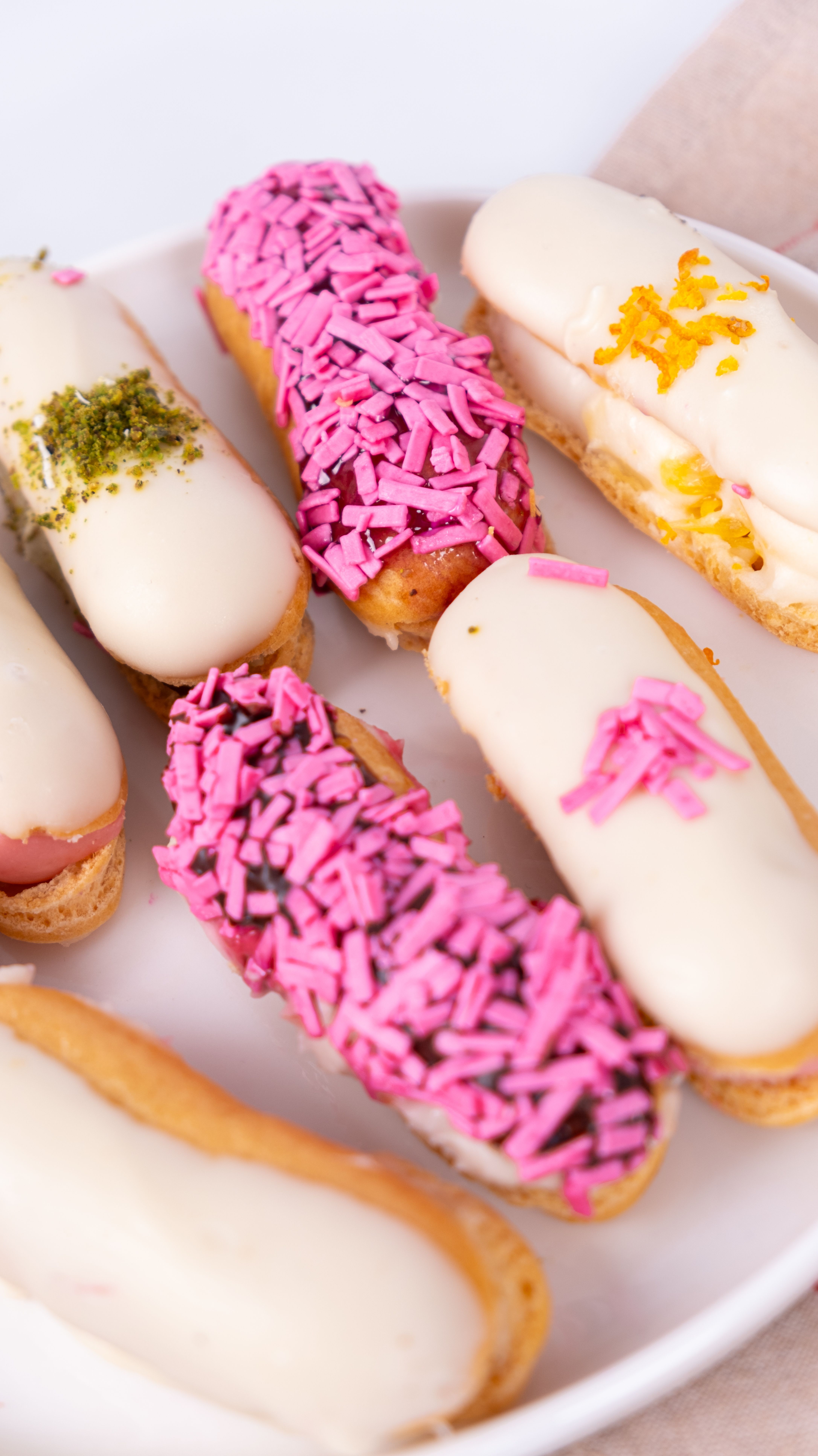 A close-up of assorted eclairs topped with white glaze and colorful sprinkles on a plate.