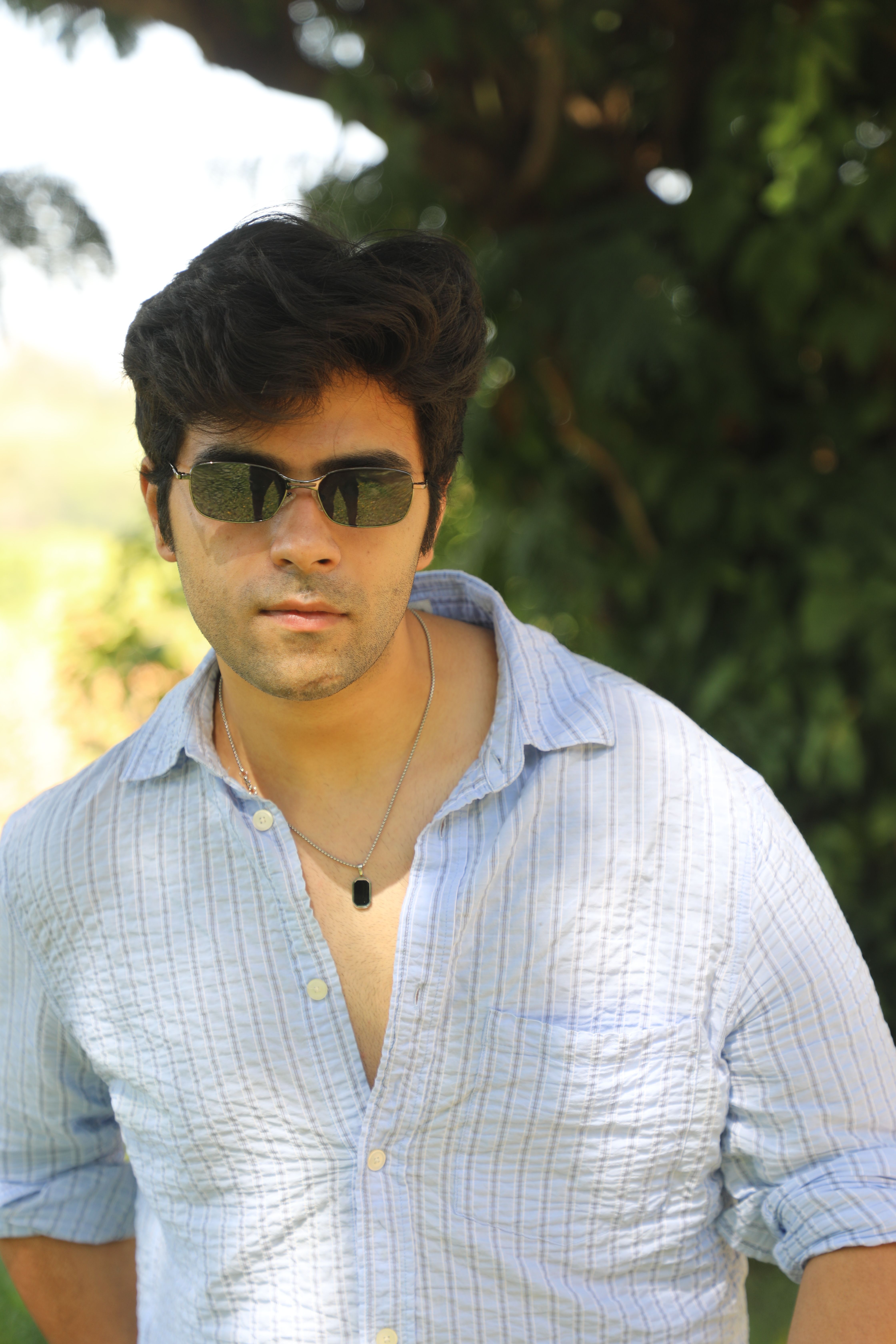 A man wearing sunglasses poses outdoors, exuding confidence in a casual shirt.