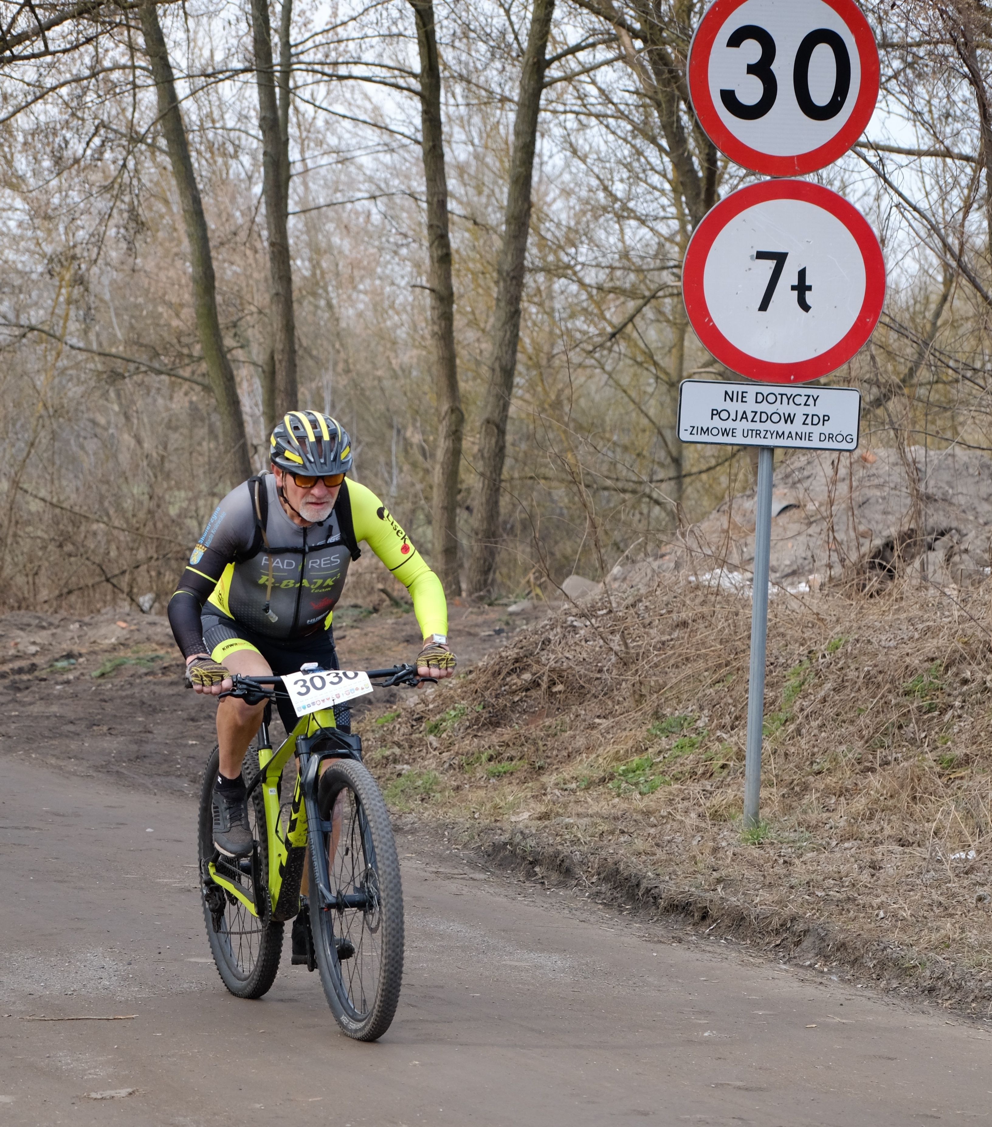 Cyclist in a racing outfit competes on a dirt path beside speed limit signs in an outdoor setting.