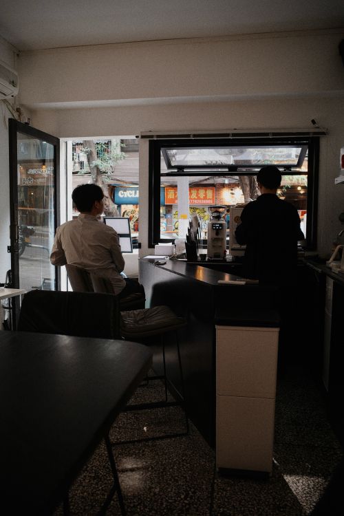 Free Two people in a cozy coffee shop, one seated and one working behind the counter. Stock Photo