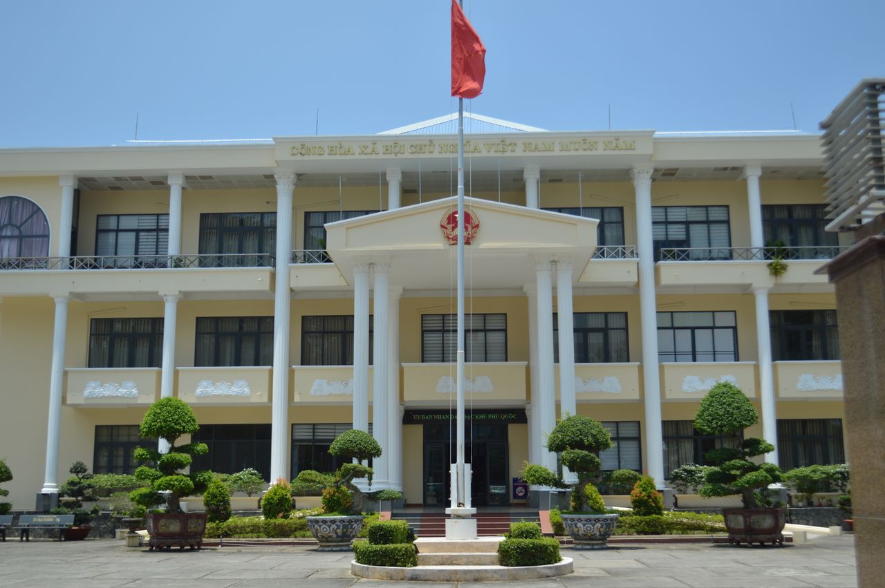 Front view of a government building in Phu Quoc, Vietnam, with a red flag and decorative plants.