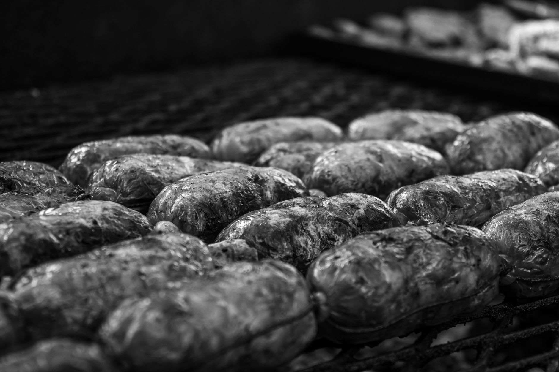 Black and white close-up image of sausages being grilled over an outdoor barbecue pit.