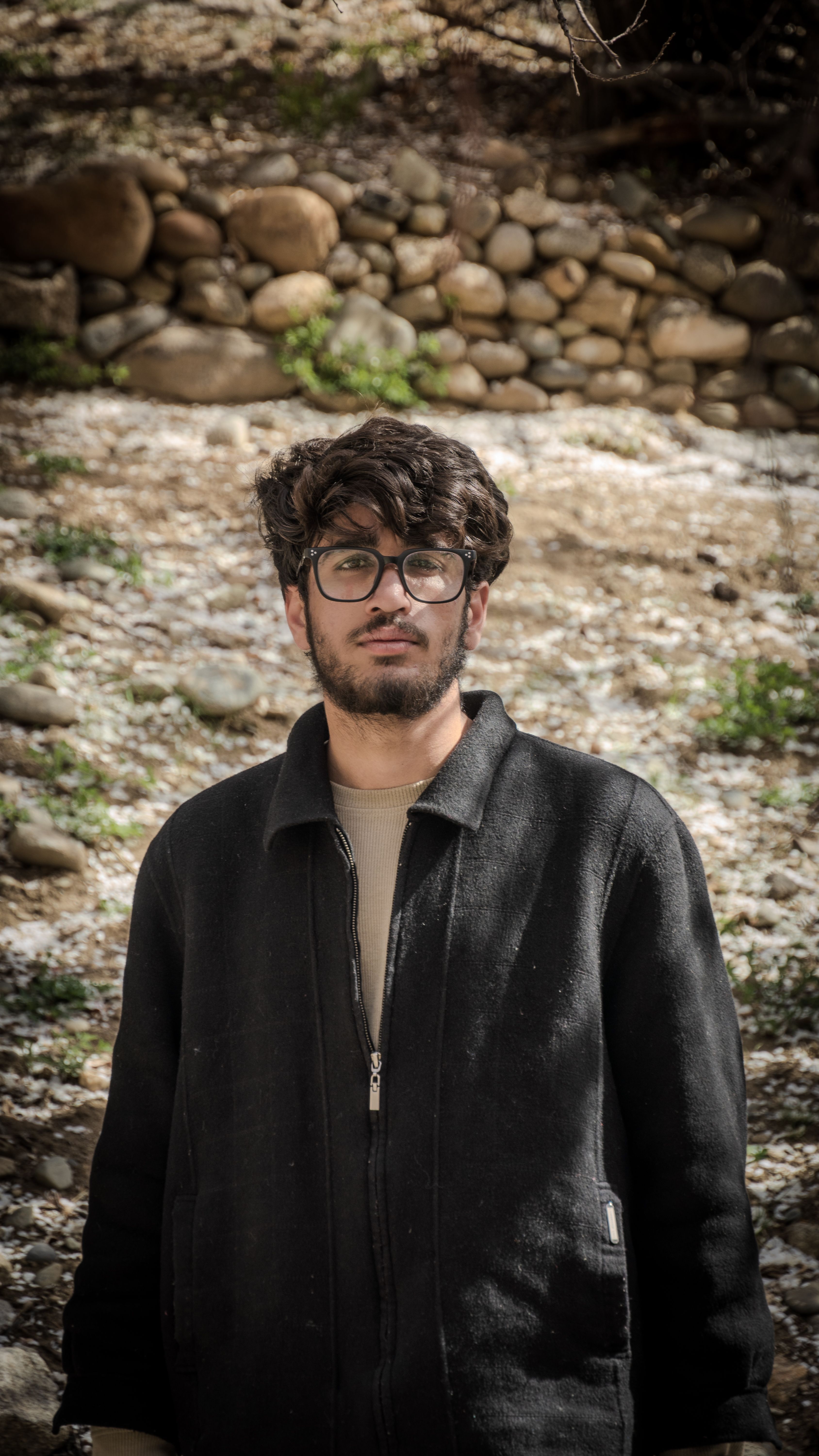 A young man with glasses standing outdoors against a rocky background on a sunny day.