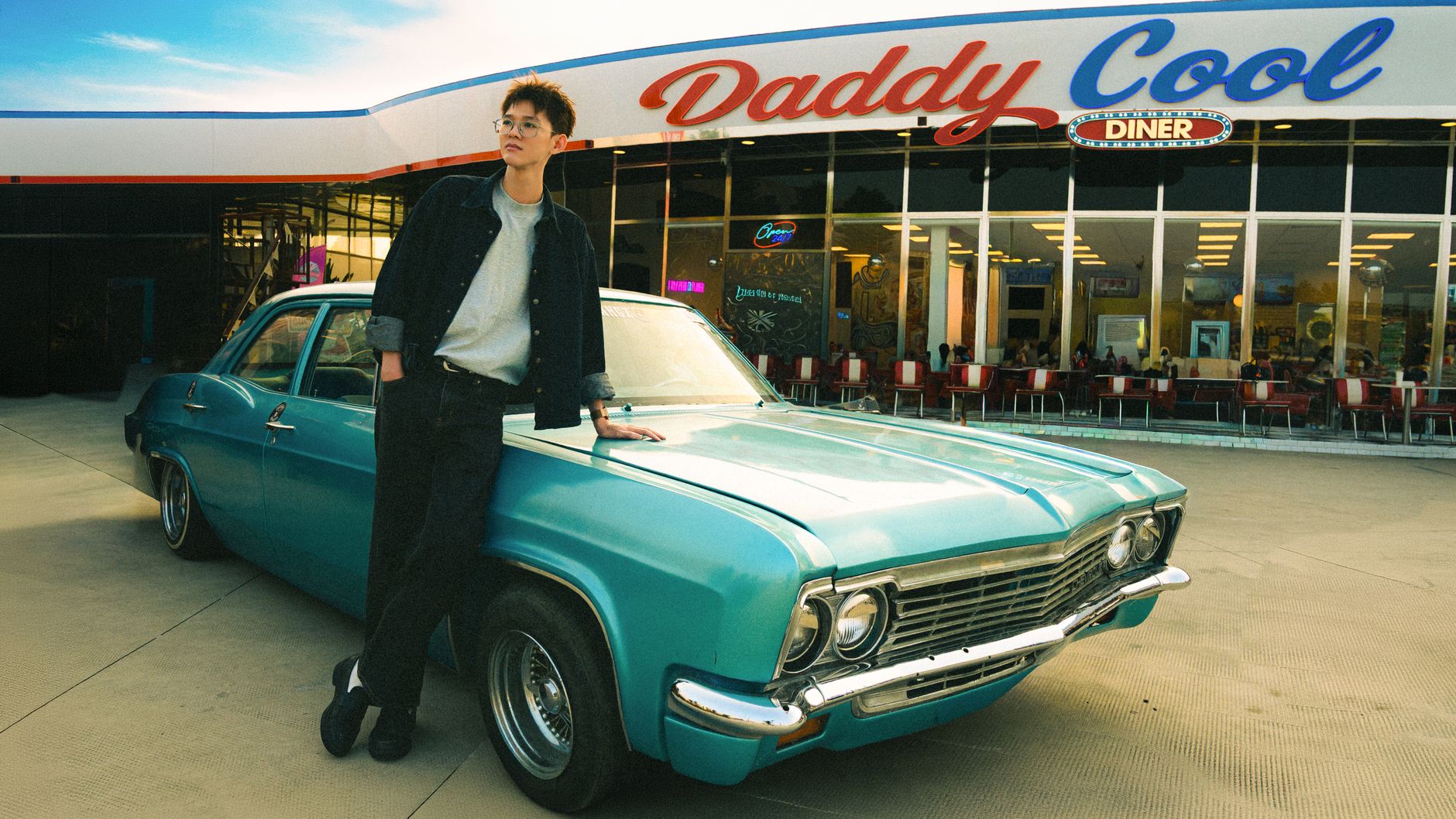 Young man leaning on classic blue vintage car in front of retro diner.