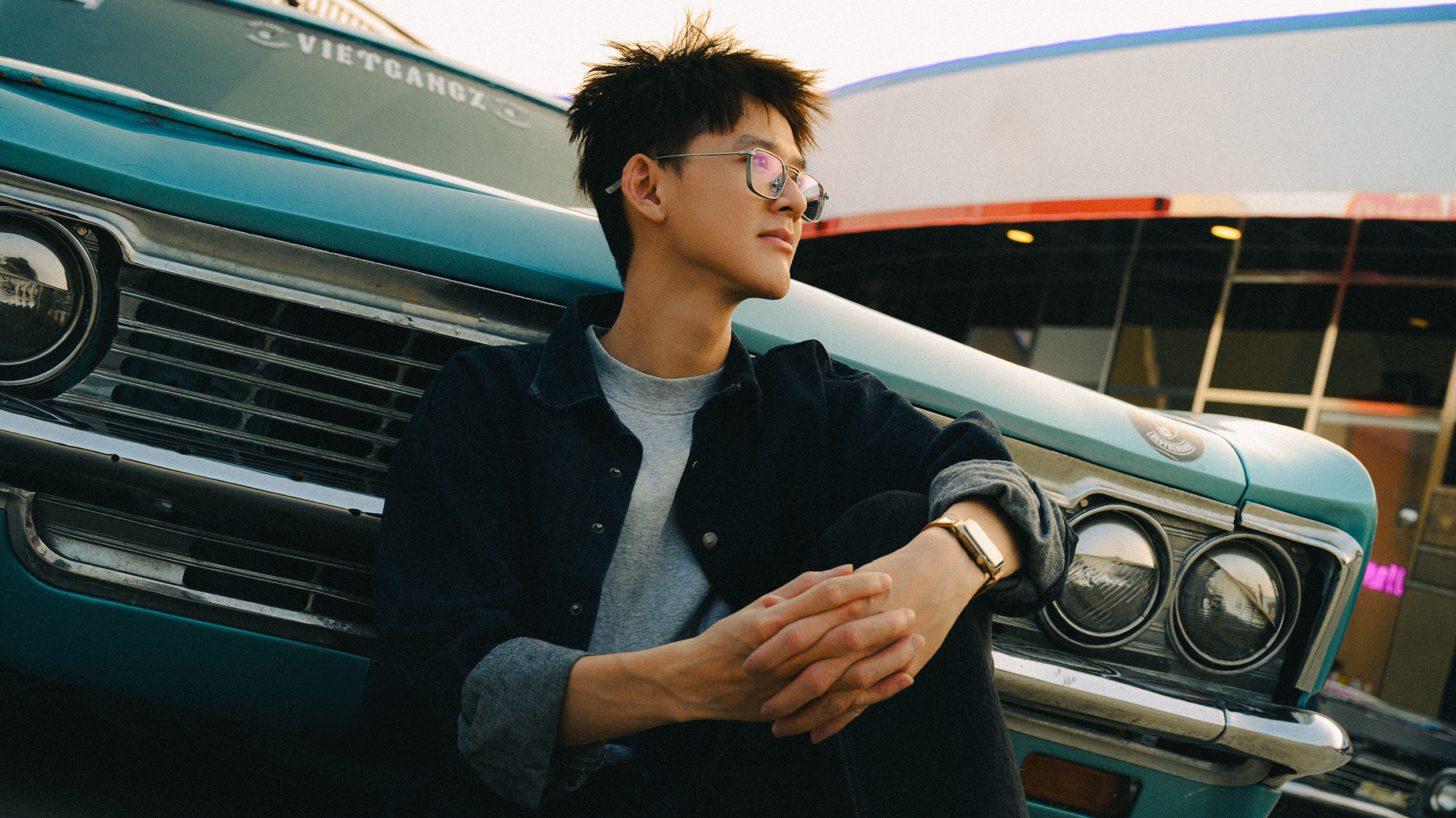 Young man sitting by turquoise classic car outdoors, reflective and relaxed.