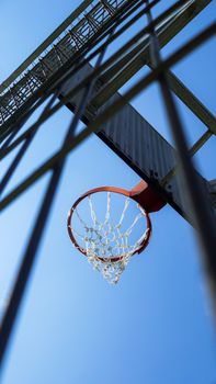 A low-angle view of a basketball hoop outdoors, framed by a fence against a clear blue sky.