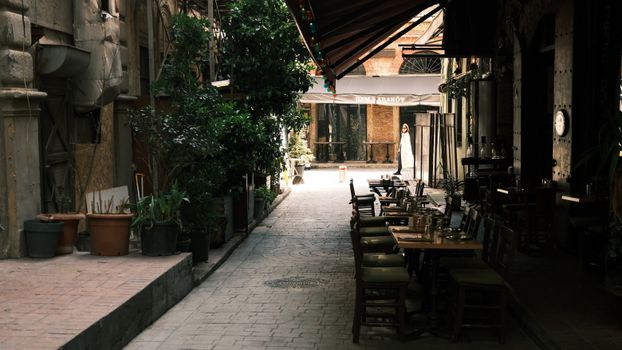 Cozy alleyway with outdoor cafe seating, lush greenery, and a solitary pedestrian.