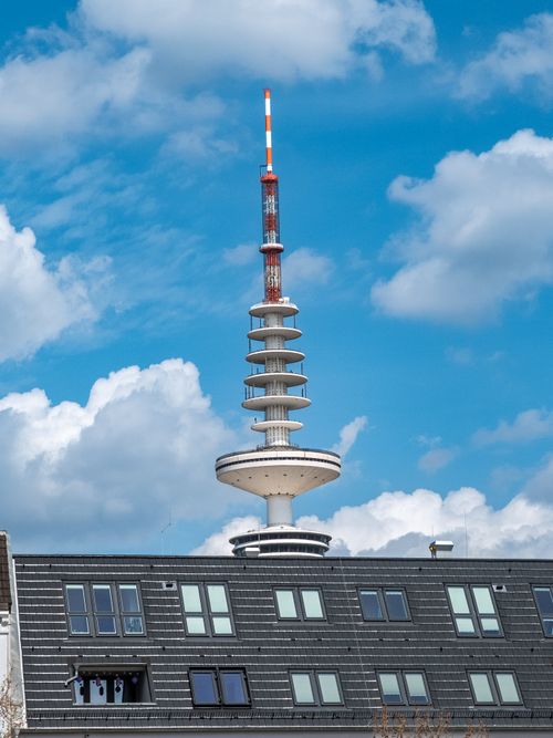 Free A television tower stands tall above urban buildings under a dramatic blue sky with clouds. Stock Photo