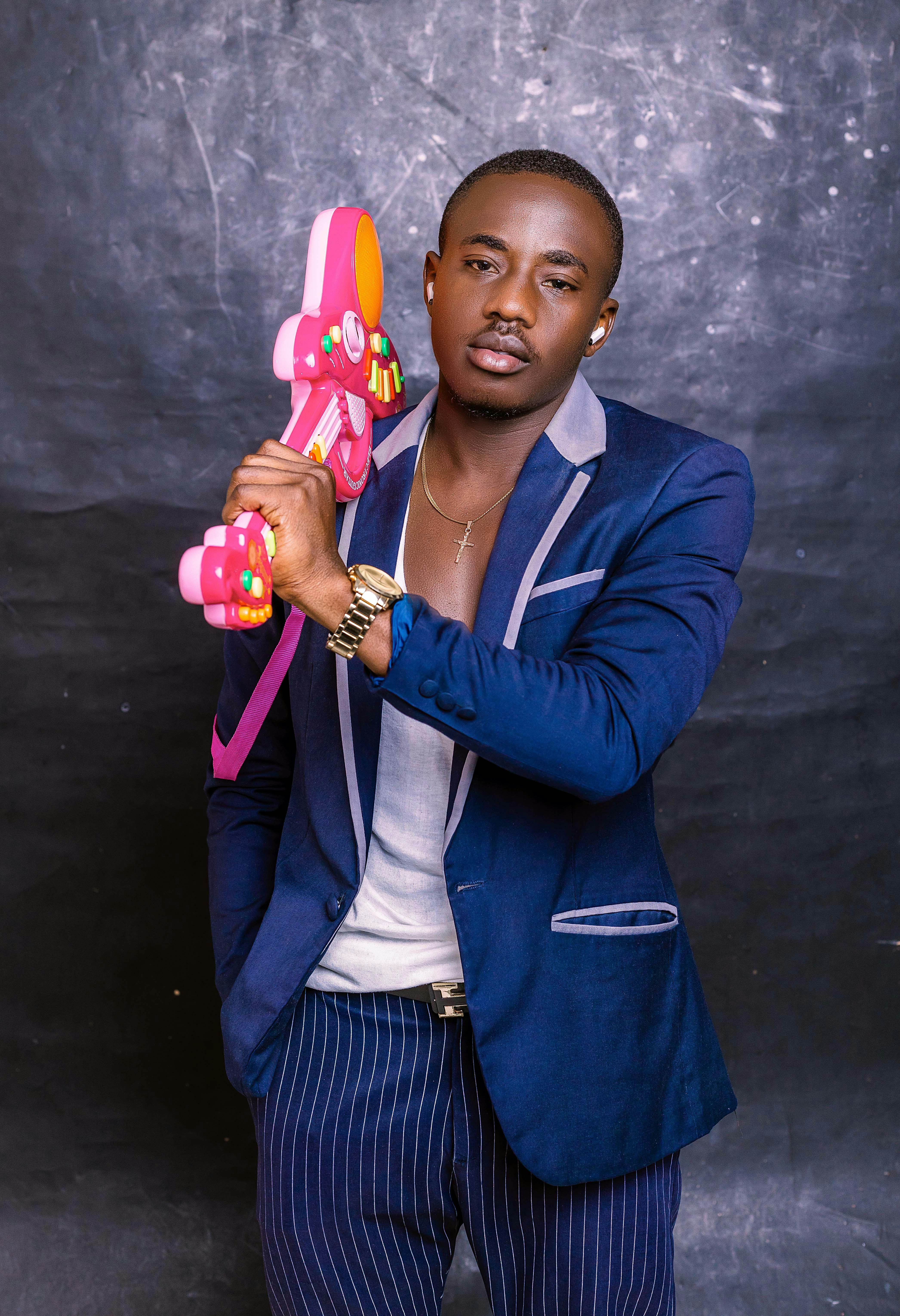 Fashionable man in blue suit posing with a pink toy guitar against a dark background.