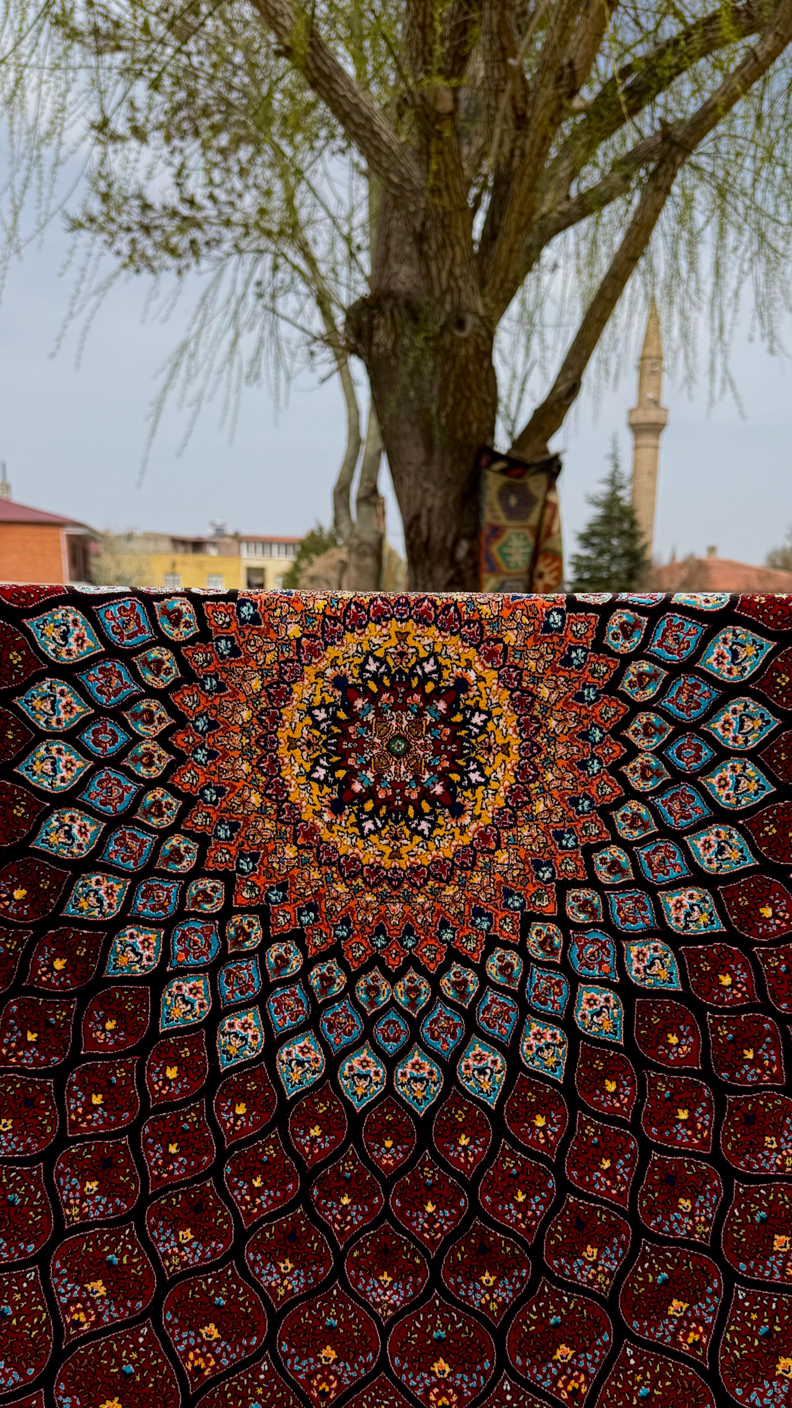 Brightly colored tapestry with intricate patterns held outdoors near tree and tower.