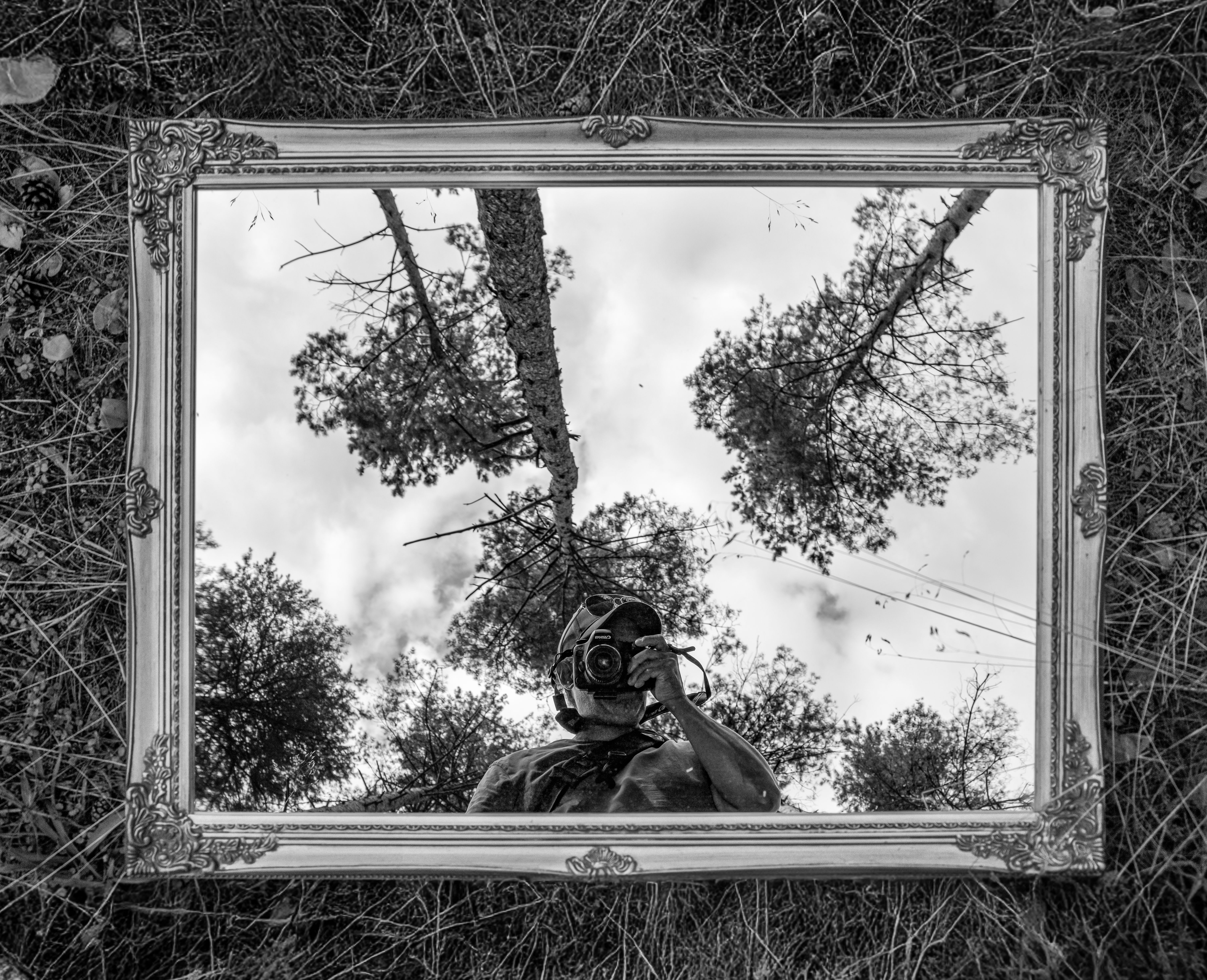 Black and white photo of a photographer's reflection in a framed mirror outdoors.
