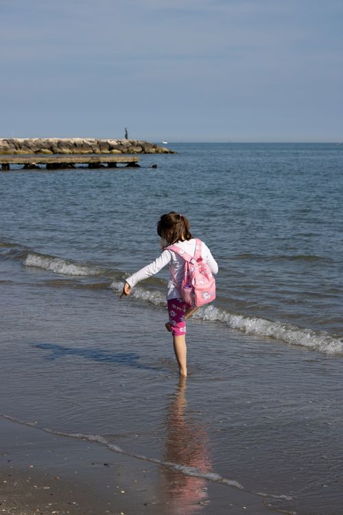 Free A young girl with a pink backpack enjoys a day at the beach, stepping into the water. Stock Photo