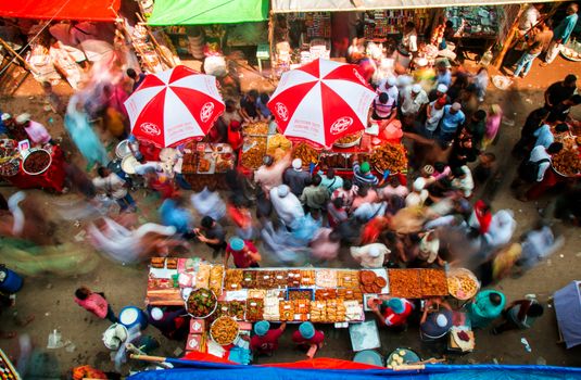 A bustling outdoor market with colorful stalls and moving crowds viewed from above.