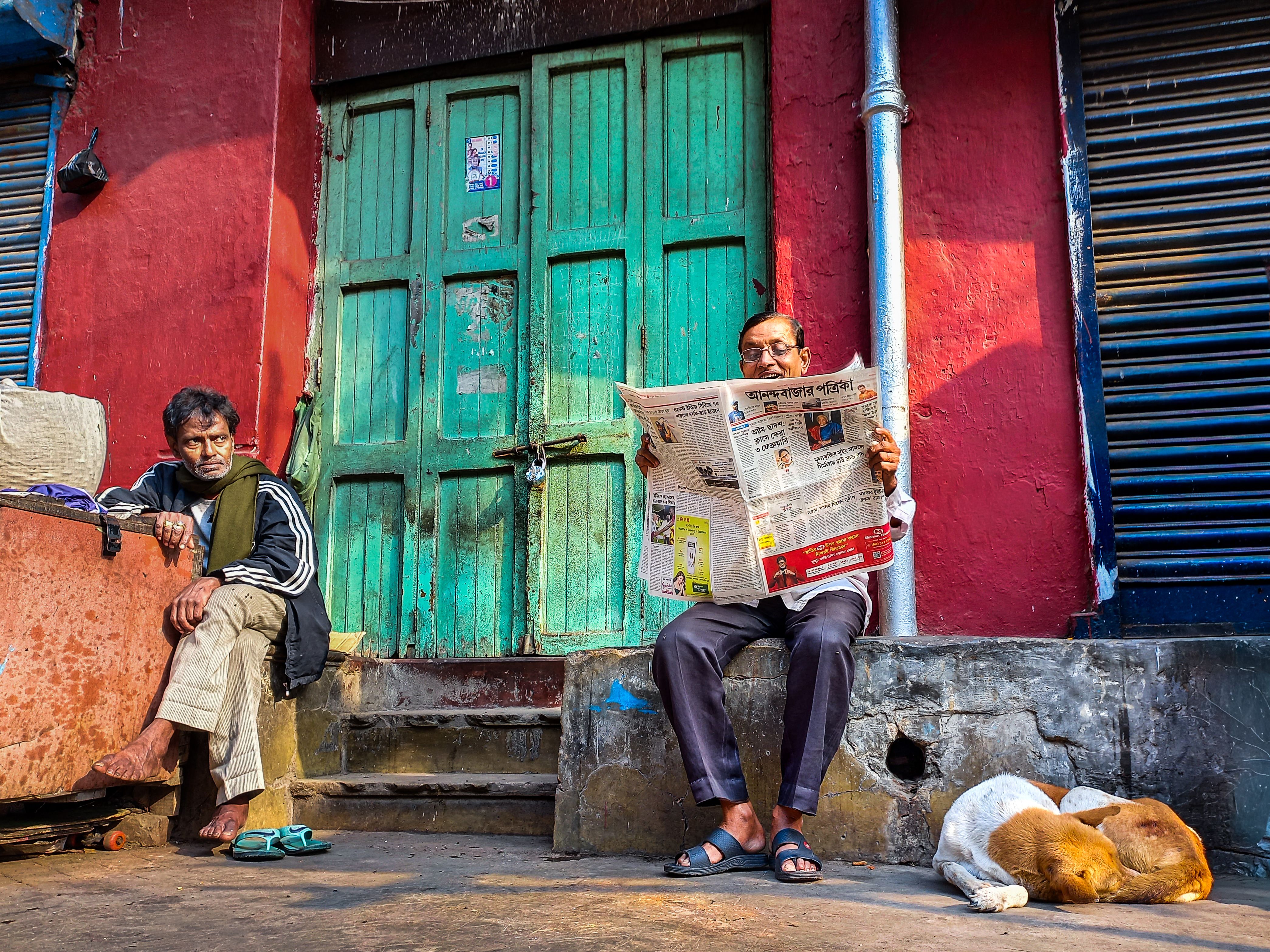A man reads a newspaper on a vibrant street while another man and a dog relax nearby.