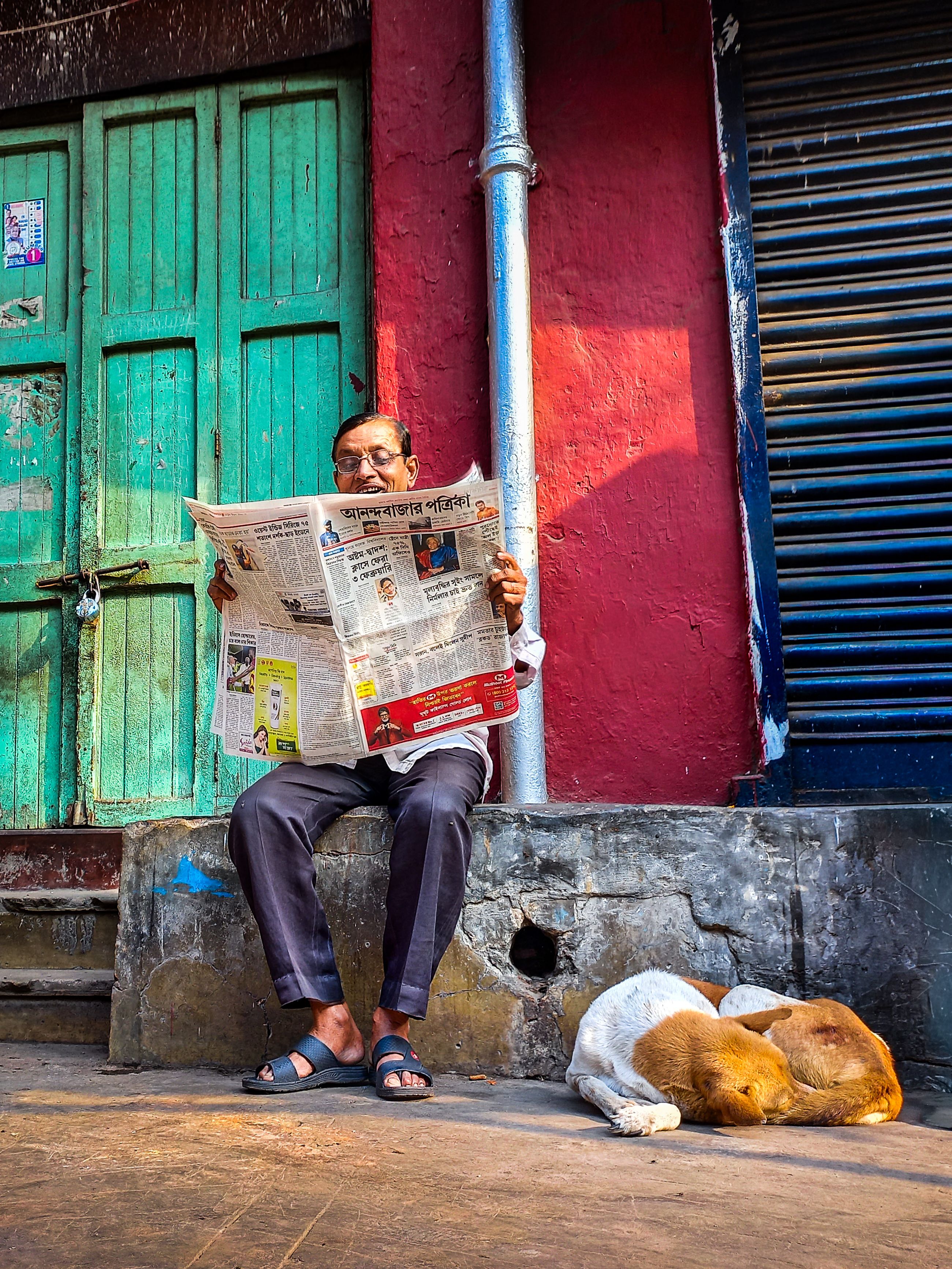 A man reads a newspaper on a street bench with a dog sleeping nearby in a vibrant urban scene.