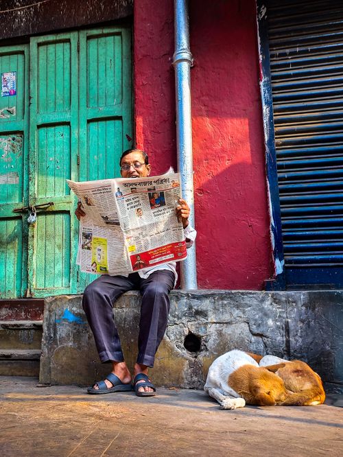 Free A man reads a newspaper on a street bench with a dog sleeping nearby in a vibrant urban scene. Stock Photo