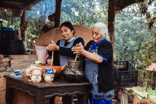 Two elderly women prepare traditional food outdoors in a rustic kitchen surrounded by nature.