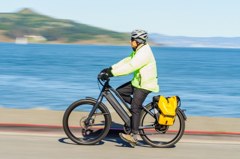 A person in neon gear cycles an e-bike with a yellow pannier on a seaside road.