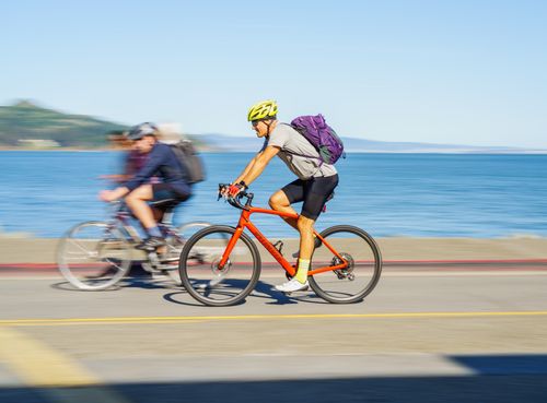 Free Two cyclists speed along a scenic coastal road, enjoying the vibrant outdoor summer weather. Stock Photo