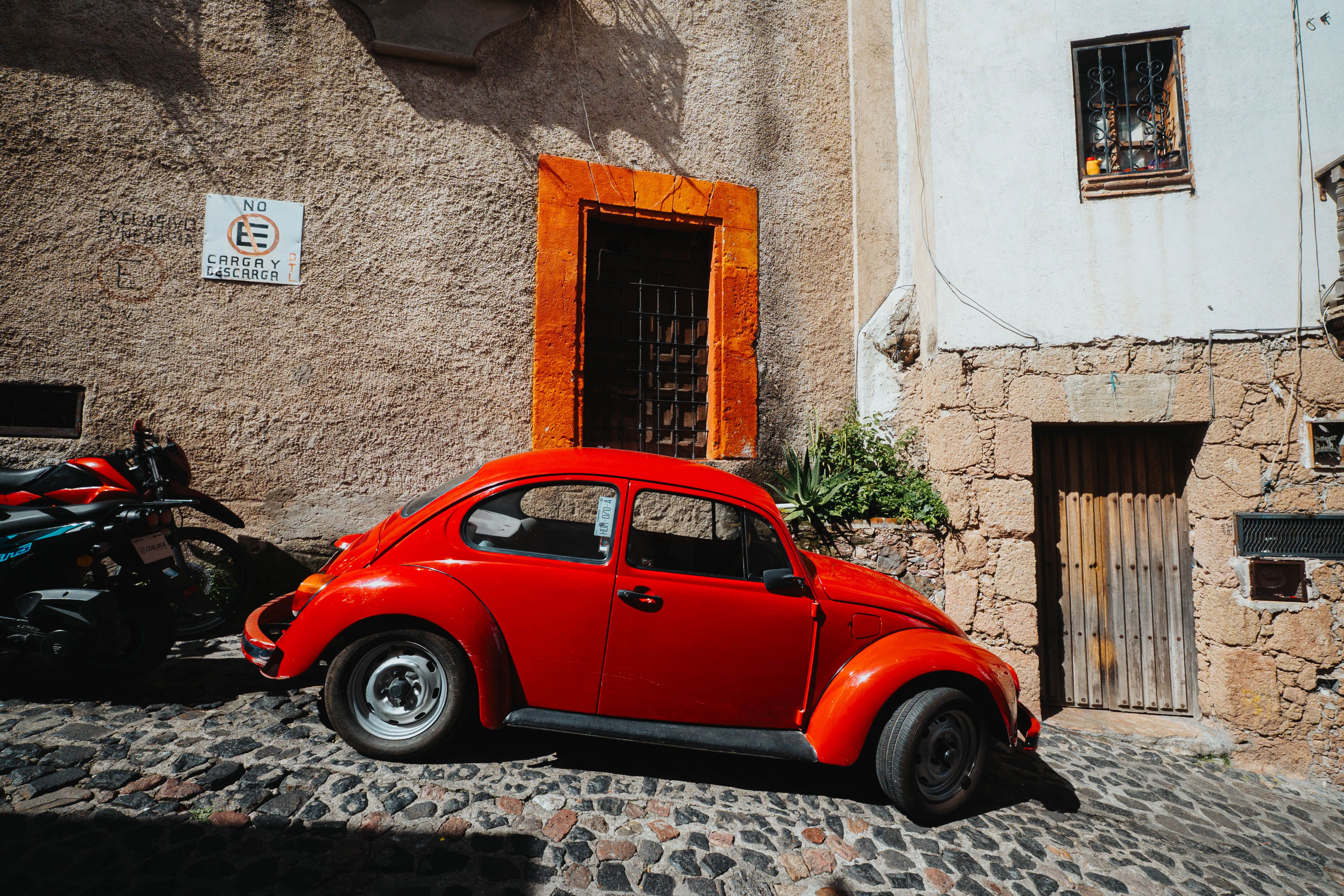 A classic red Beetle car parked on a cobblestone street beside a rustic building under bright sunlight.