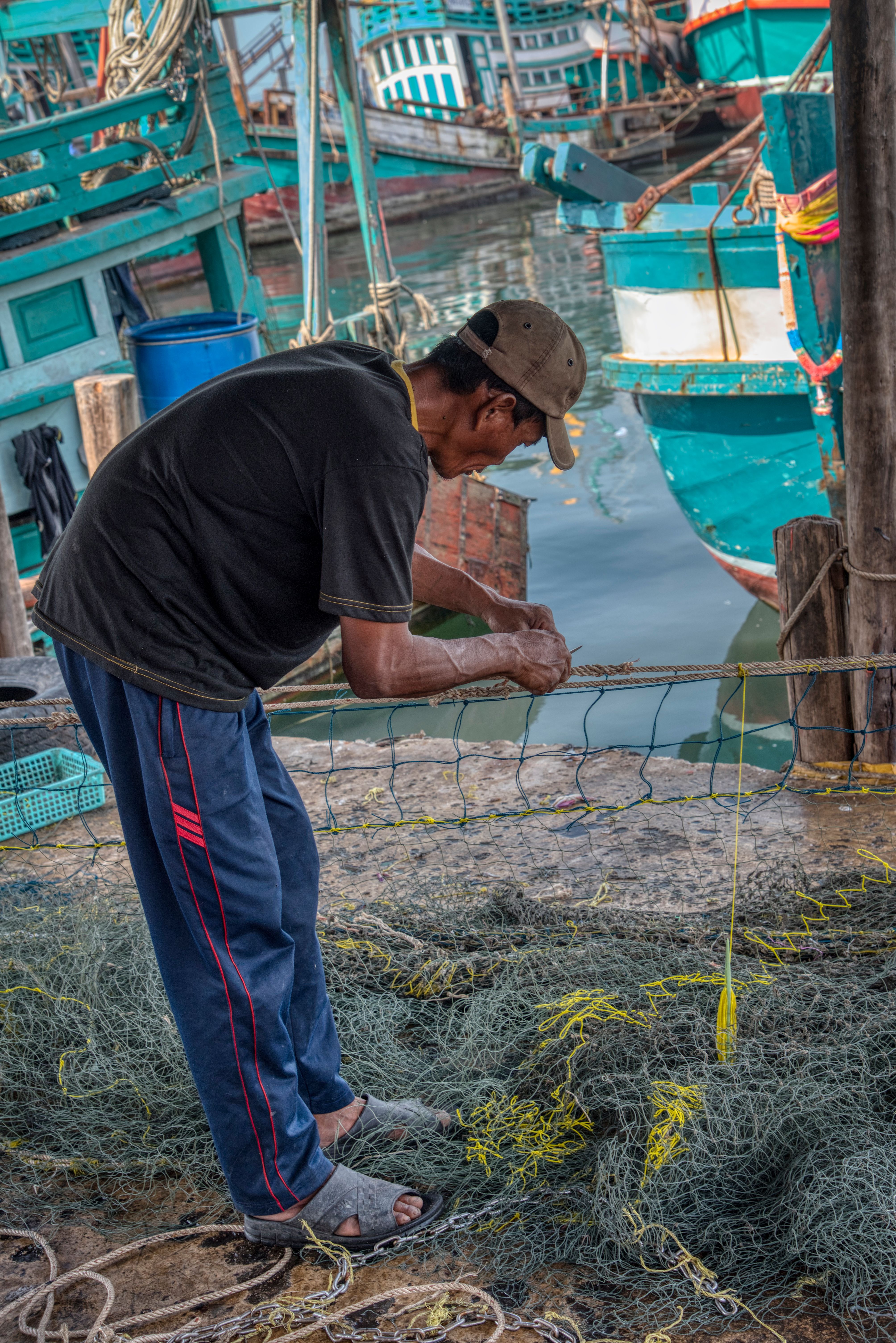 An adult fisherman mending fishing nets at an Asian dock with boats in the background.